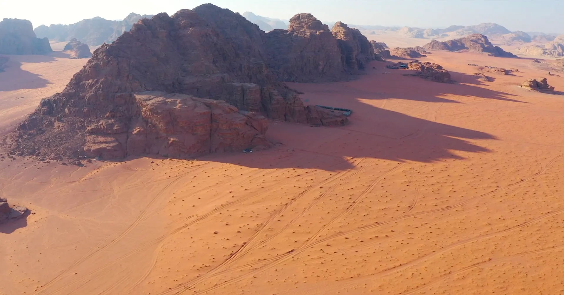 This image shows a desert landscape with large rock formations in a sandy terrain. It appears to be sunny, with shadows cast by the rocks. The landscape might resemble environments like Wadi Rum or similar desert areas.