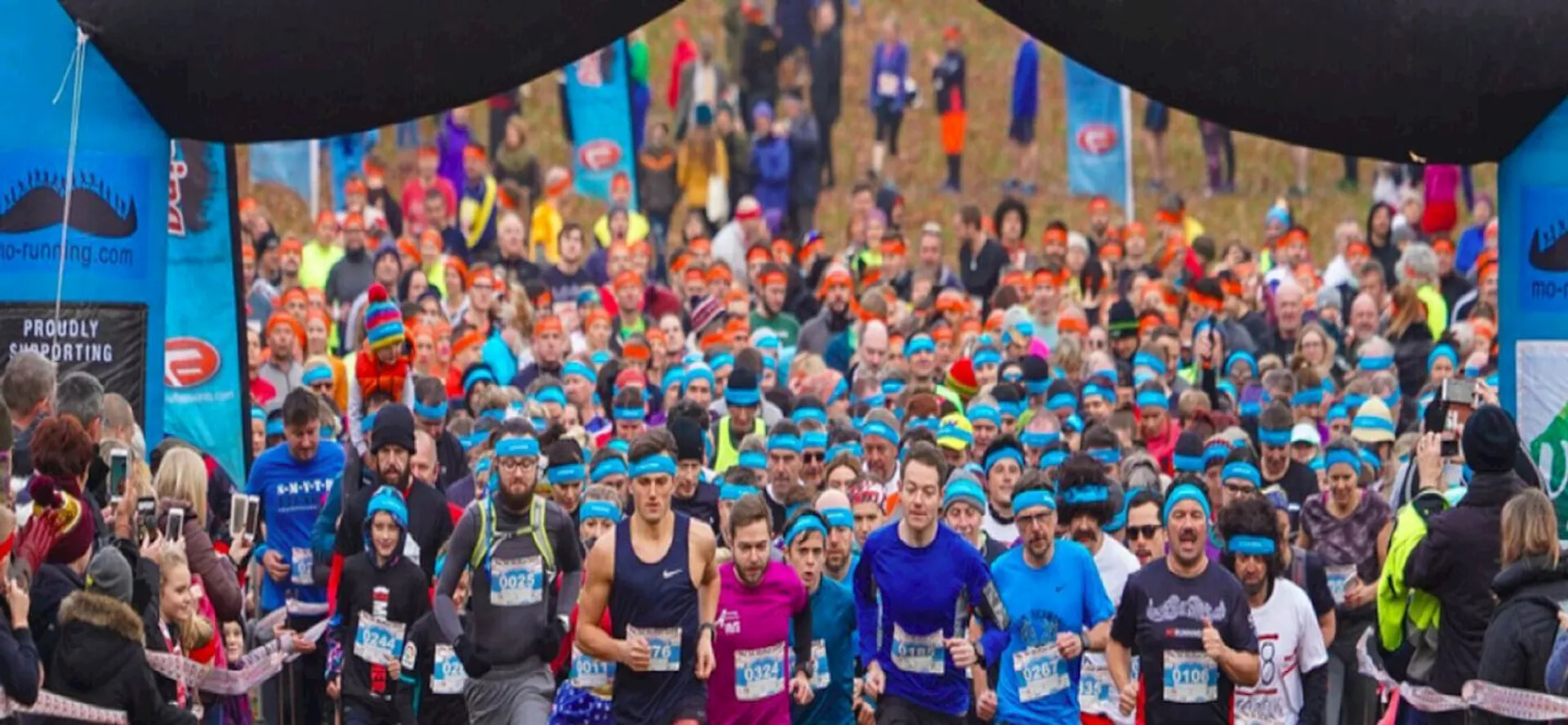 The image shows a large group of runners participating in a race. They are passing under a starting or finishing arch. Many of the runners are wearing blue headbands, and there is a crowd of spectators around them. The scene is colorful and energetic.