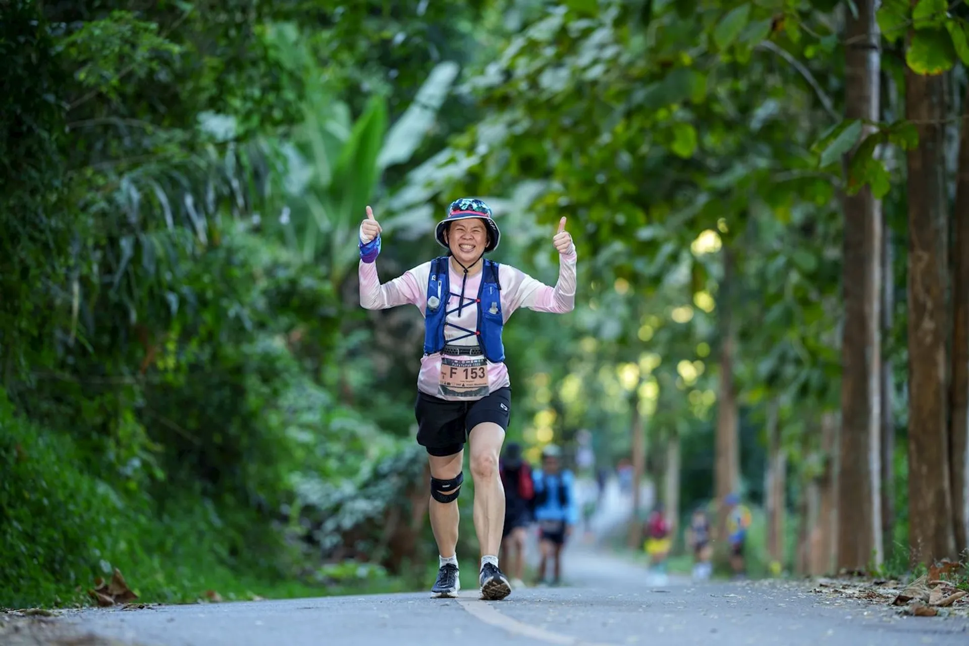 The image shows a person running on a road surrounded by greenery. The runner is giving thumbs up, wearing a bucket hat, a hydration vest, and has a race bib. Other runners can be seen in the background. It appears to be an outdoor running event or marathon.