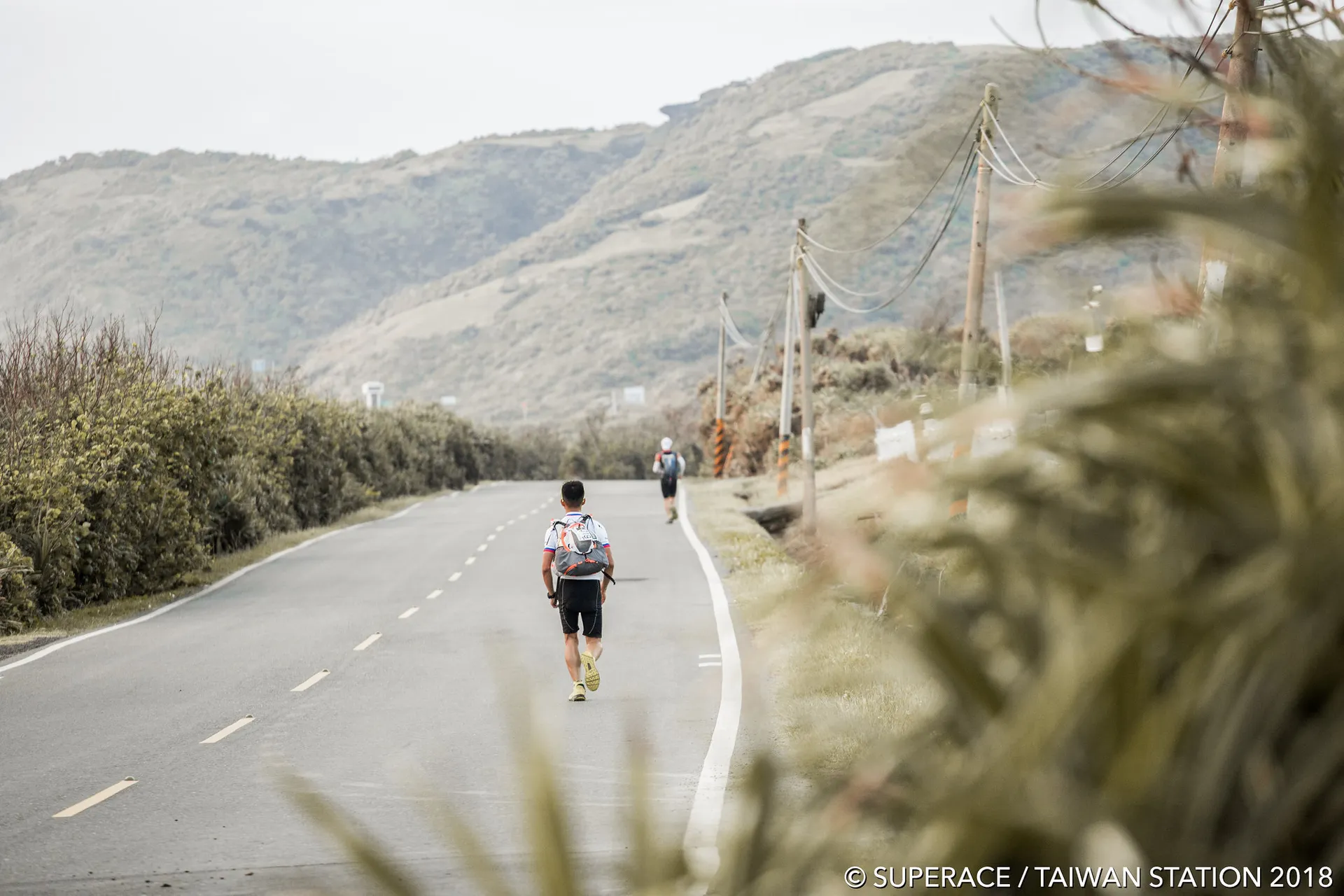 The image depicts a runner on a road with a scenic background. The runner is wearing a backpack and appears to be participating in a long-distance running event, possibly an ultra-marathon given the setting and the gear. The road bends gently to the left, and there are lush green hills in the distance, suggesting this could be a rural or mountainous area. There's another participant visible further up the road. The vegetation to the right indicates a windy environment or coastal area, as the plants are leaning to one side. This suggests that the runners might be facing some windy conditions. The watermark indicates that this is from an event called SUPERACE, located in Taiwan, and the photo was taken in 2018.