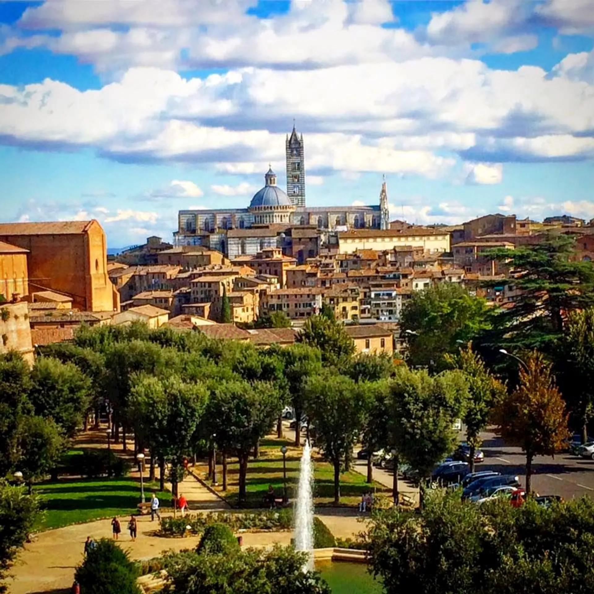 The image you've provided features a scenic view of a historic cityscape with a prominent cathedral rising above the other buildings, showcasing a tall bell tower and a large dome. In the foreground, there's a well-manicured park with a fountain, trees neatly lined up, and people walking along pathways. The architecture suggests a European city, possibly Italian given the style of the dome and tower, reminiscent of Renaissance architecture. The buildings are densely packed, showing off warm colors and terracotta roofs, typical of many historic Italian town centers. The sky is partly clouded, suggesting a pleasant and peaceful day, ideal for sightseeing or relaxing in the park.