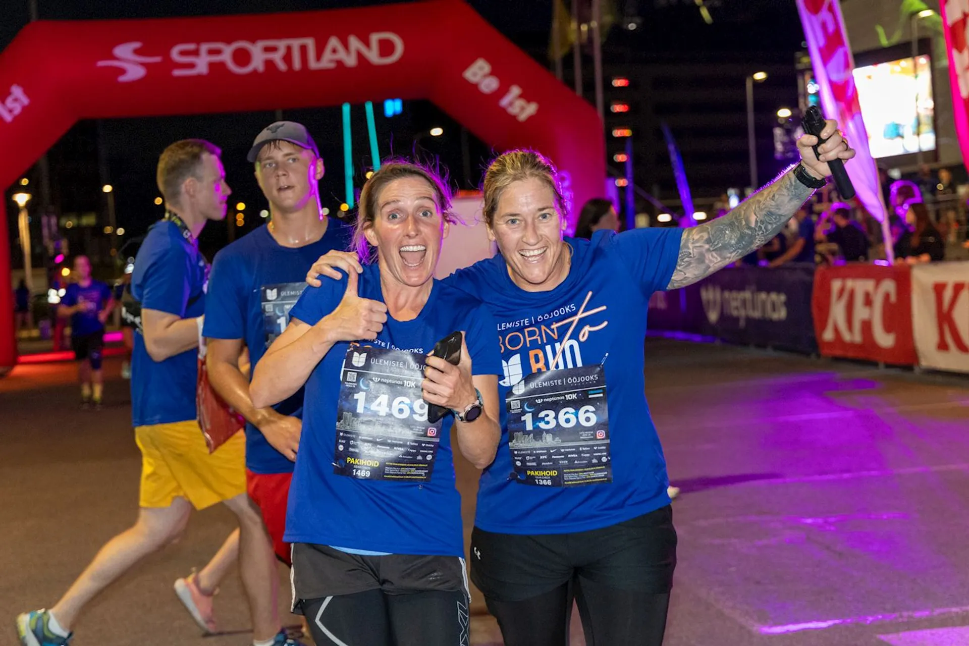 In this image, there are four individuals, presumably athletes, participating in a night running event. They appear to be crossing the finish line, as suggested by the inflatable arch with the word "FINISH" and various sponsor logos on it. Two women in the foreground are celebrating their accomplishment; one of them holds up her right arm in a gesture of triumph, and both are smiling broadly and displaying their race bibs, indicating their participation in the event. The runners are wearing matching blue event T-shirts, suggesting that this may be a team or group run. The atmosphere of the event is festive and lively, as seen from the bright lighting and the presence of multiple participants.