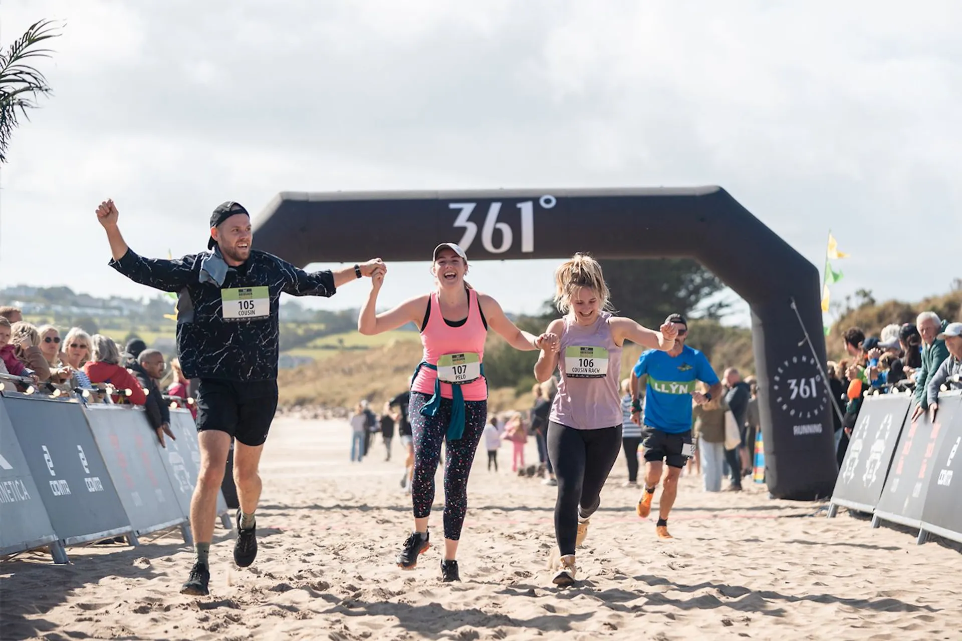 The image shows a group of people running towards a finish line on a sandy surface, likely a beach. They appear to be participating in a race or marathon event. There’s an inflatable arch in the background with "361°" on it, which could be the branding for the event. Participants and spectators are visible in the background, adding to the lively atmosphere.