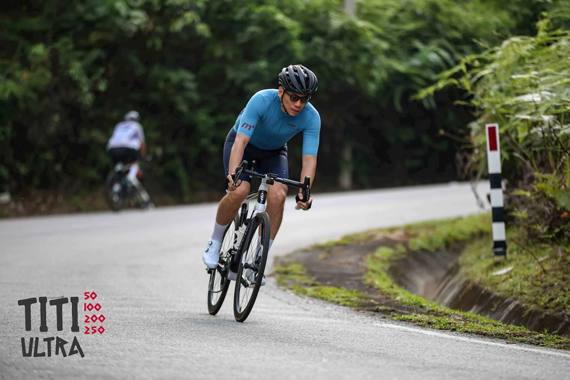 The image shows a cyclist wearing a helmet and blue cycling jersey, riding on a road with greenery in the background. There is another cyclist visible in the distance. The text on the image reads "TIT ULTRA" with numbers 50, 100, 200, and 250, which may refer to distances in a race or event.
