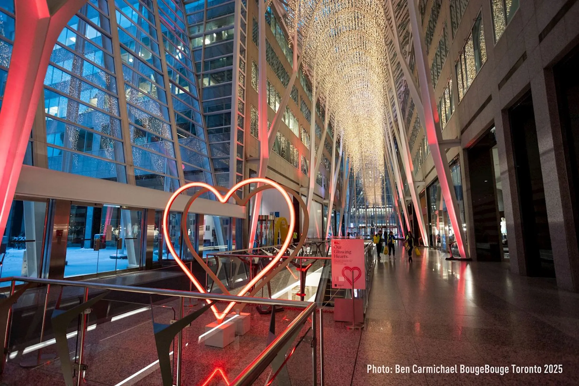 This image shows an interior space with a large, illuminated heart sculpture in the foreground. The area has tall, arching metallic structures and glass walls, giving it a modern and open feel. The ceiling is adorned with decorative lighting, and people can be seen in the background. This is likely a public or commercial space.