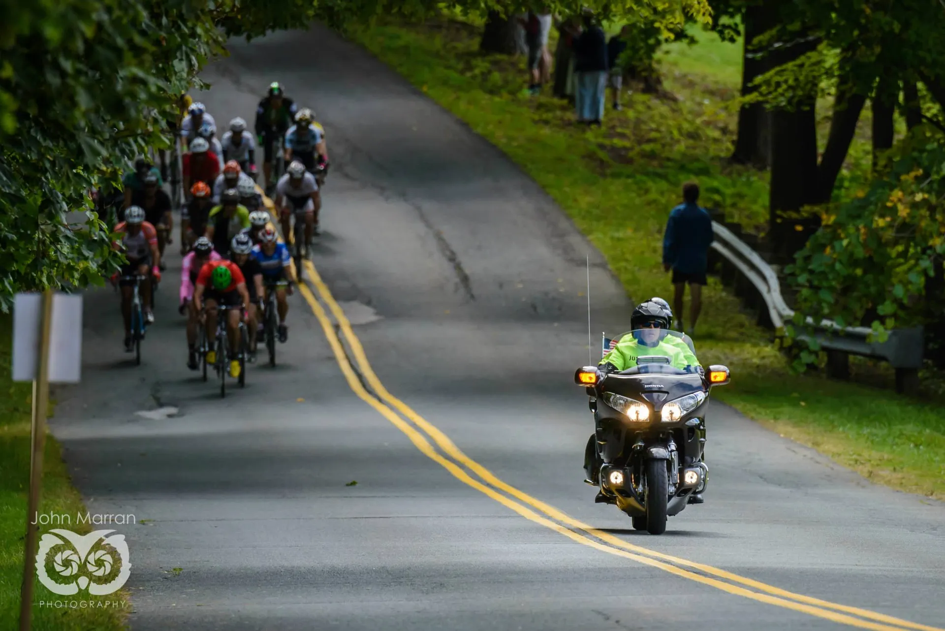 The image shows a group of cyclists riding on a road during what appears to be a cycling event. In the foreground, there is a motorcyclist driving in the same direction, likely providing support or escort for the cycling event. The motorcyclist is wearing a high-visibility vest, which is common for event staff or safety personnel. The road is surrounded by green trees and grass, suggesting that this event is taking place in a park or a rural area. On the right side of the road, there are a couple of pedestrians walking on the grass, perhaps spectators or passersby. It seems to be an overcast day based on the lighting in the image.