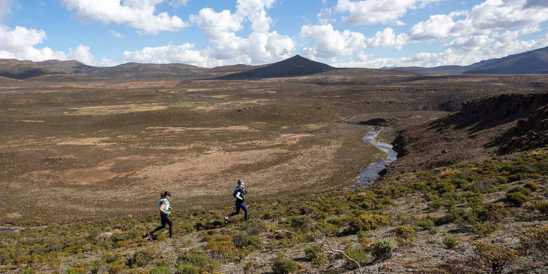 The image shows a wide landscape view with two people running on a trail in a rugged, open area. The terrain is covered with sparse vegetation and shrubs, and there's a river or stream flowing through the landscape. The sky is partly cloudy, and the distant hills or mountains create a scenic backdrop.