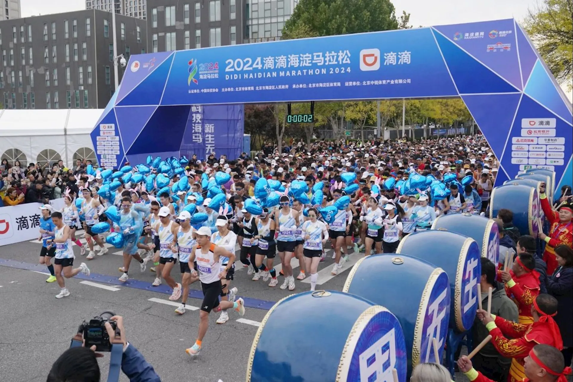 The image shows a large group of runners starting a marathon. There's a banner above the starting line with the text "2024海淀马拉松" and "Didi Haidian Marathon 2024." The race appears to be well-organized with various sponsors and event signs visible. Spectators and event staff are present on the sides, and many of the runners are carrying blue balloons.