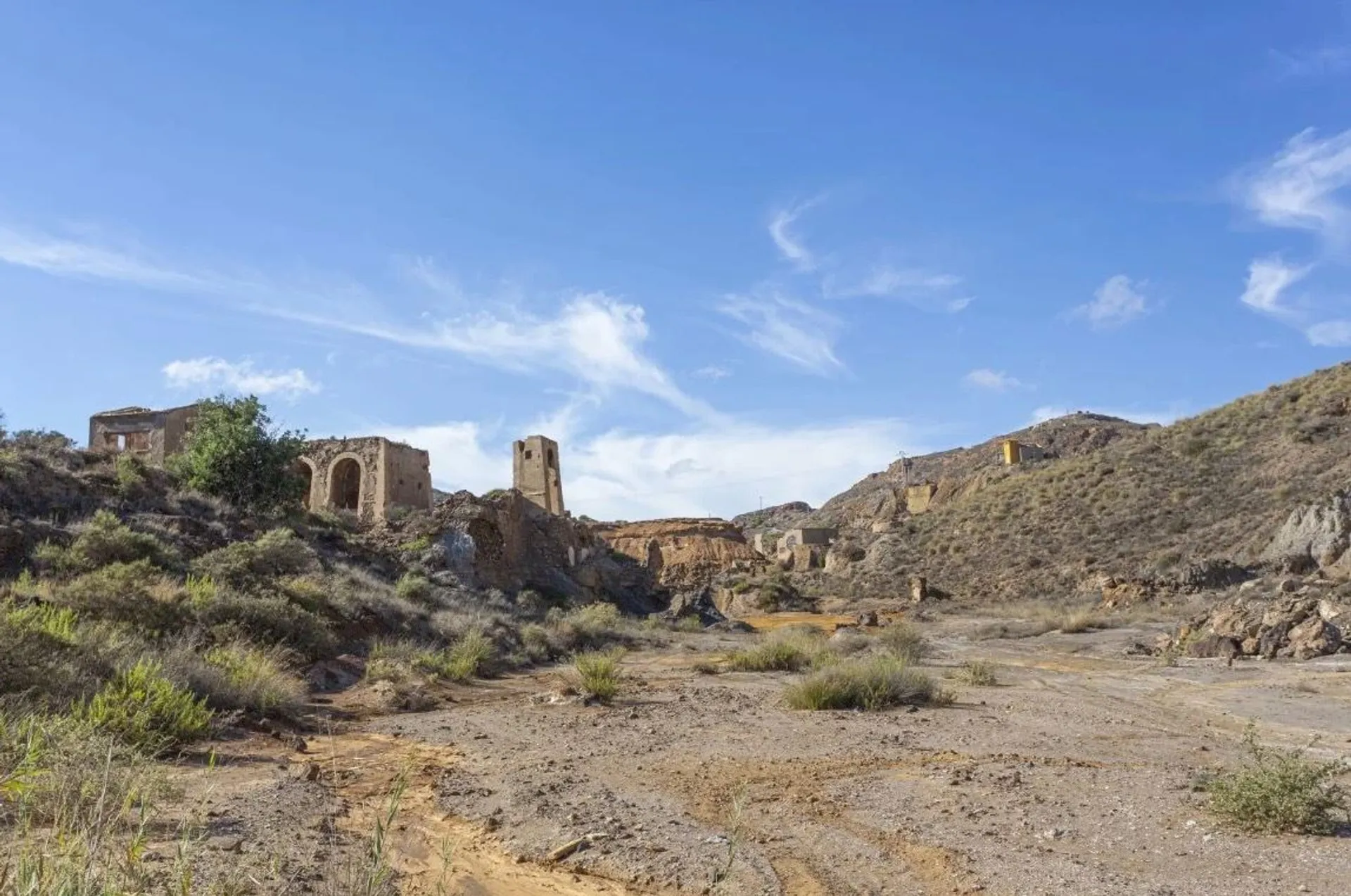 The image shows a deserted landscape with ruins of old buildings scattered across a dry, rocky terrain. There are hills in the background and a clear blue sky overhead. The area appears to be arid, with sparse vegetation and dilapidated structures, suggesting an abandoned settlement or village.
