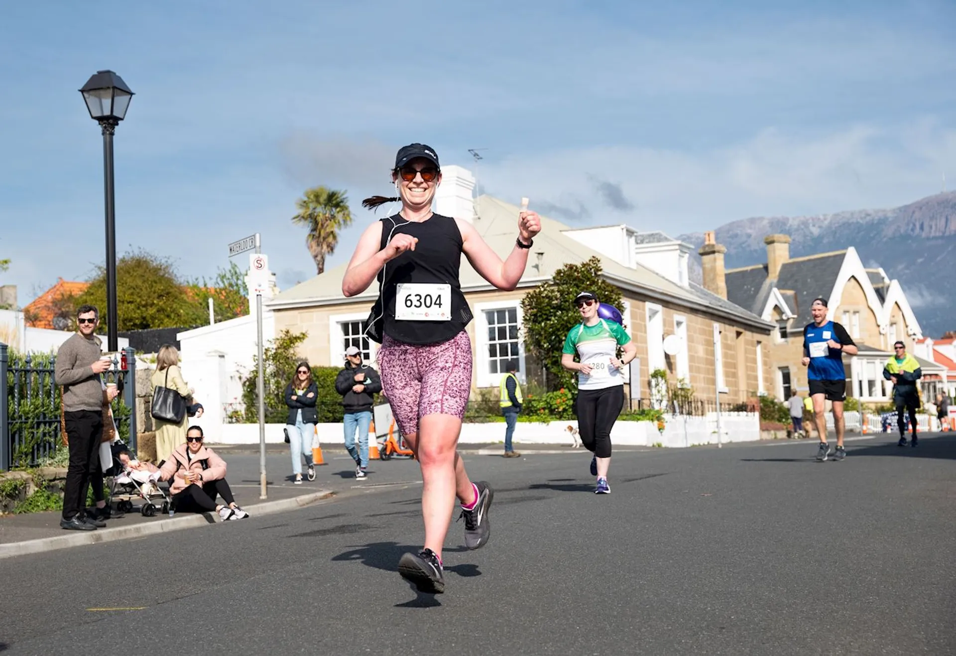 The image shows a person participating in a running event on a street. They are wearing sportswear, a cap, and a bib with the number 6304. Other runners and spectators are visible in the background, along with a few houses and a lamppost. The setting appears to be a residential area with a clear sky.