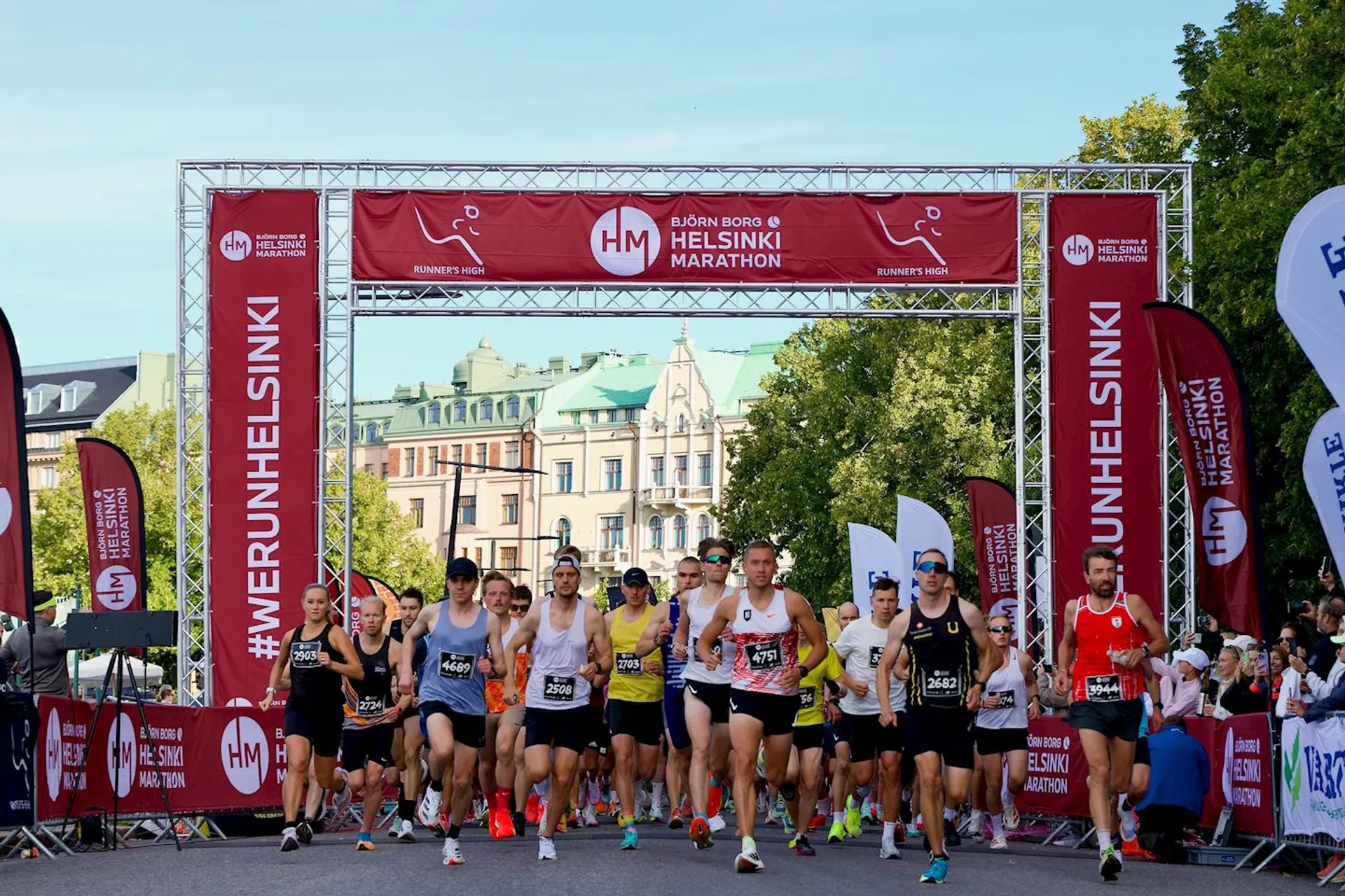 The image shows a group of runners participating in the Helsinki Marathon. They are just starting the race, as suggested by their positioning and the archway above them displaying the event's name. The street is lined with trees, and there are buildings in the background. Various banners and flags displaying event-related text are also visible.