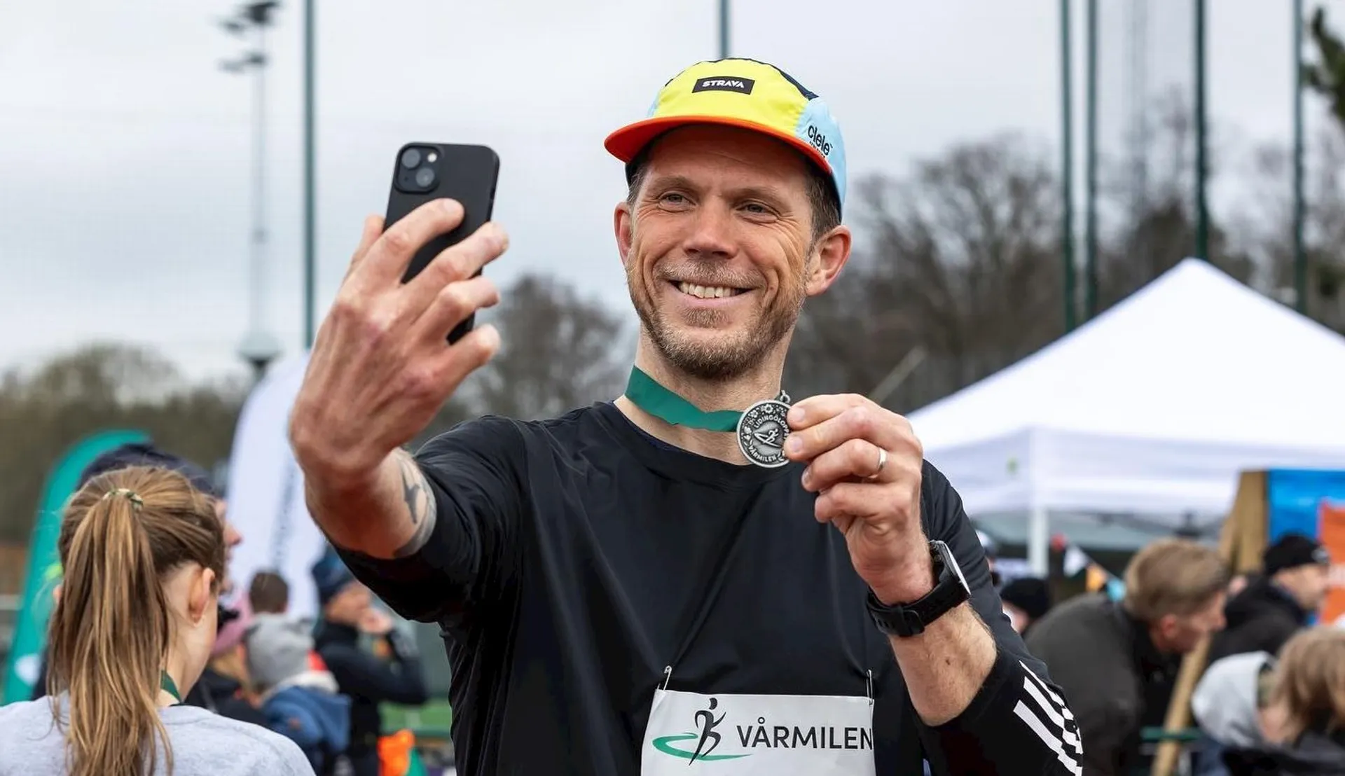 The image shows a person taking a selfie while holding a medal. They appear to be at an outdoor event with other people in the background. The person is wearing a cap and a shirt with a visible number or label that seems related to the event.