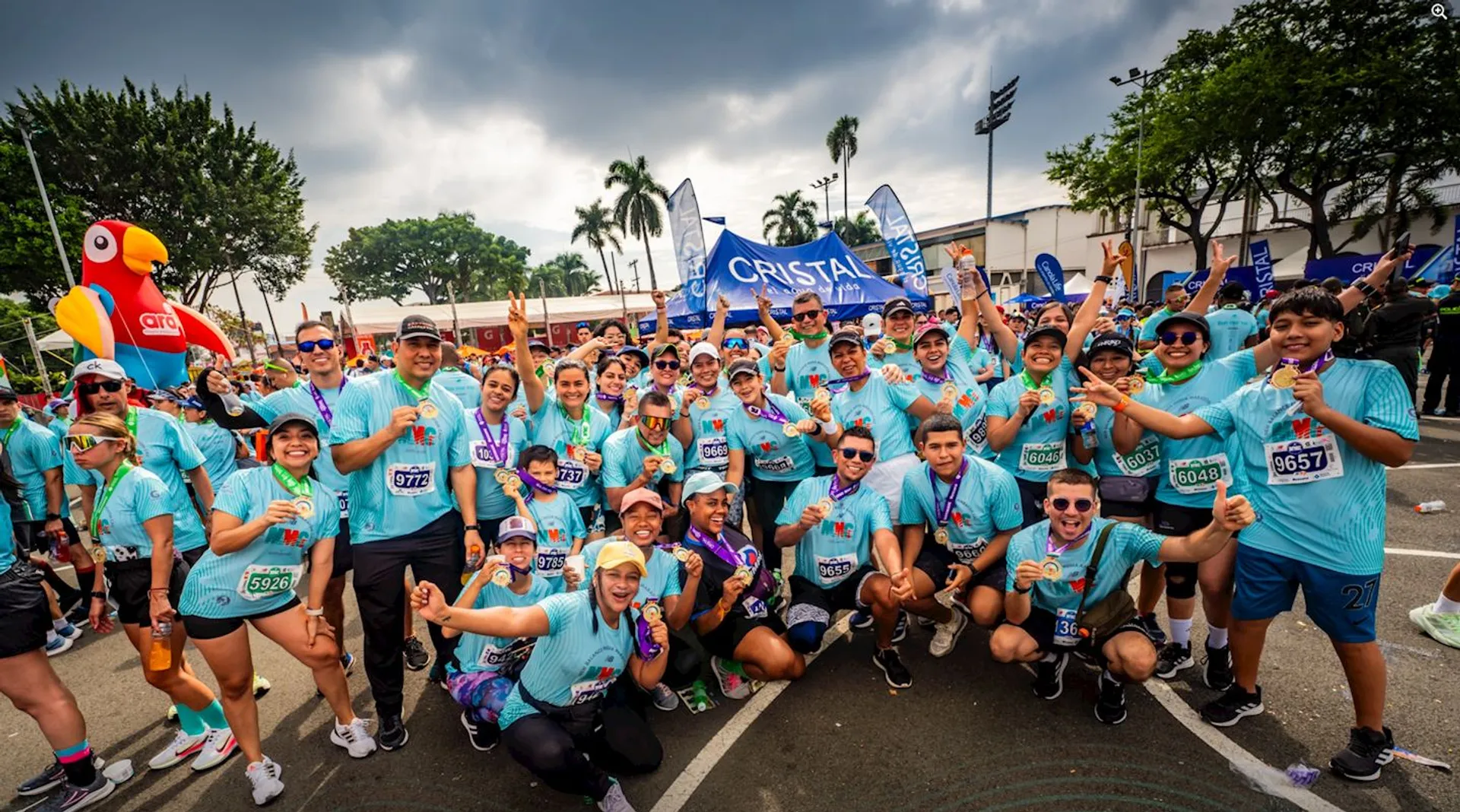 The image shows a large group of people wearing matching blue T-shirts with race bibs, indicating they participated in a race or marathon. They are celebrating, many holding medals. There's a festive atmosphere with banners and a large bird mascot in the background.
