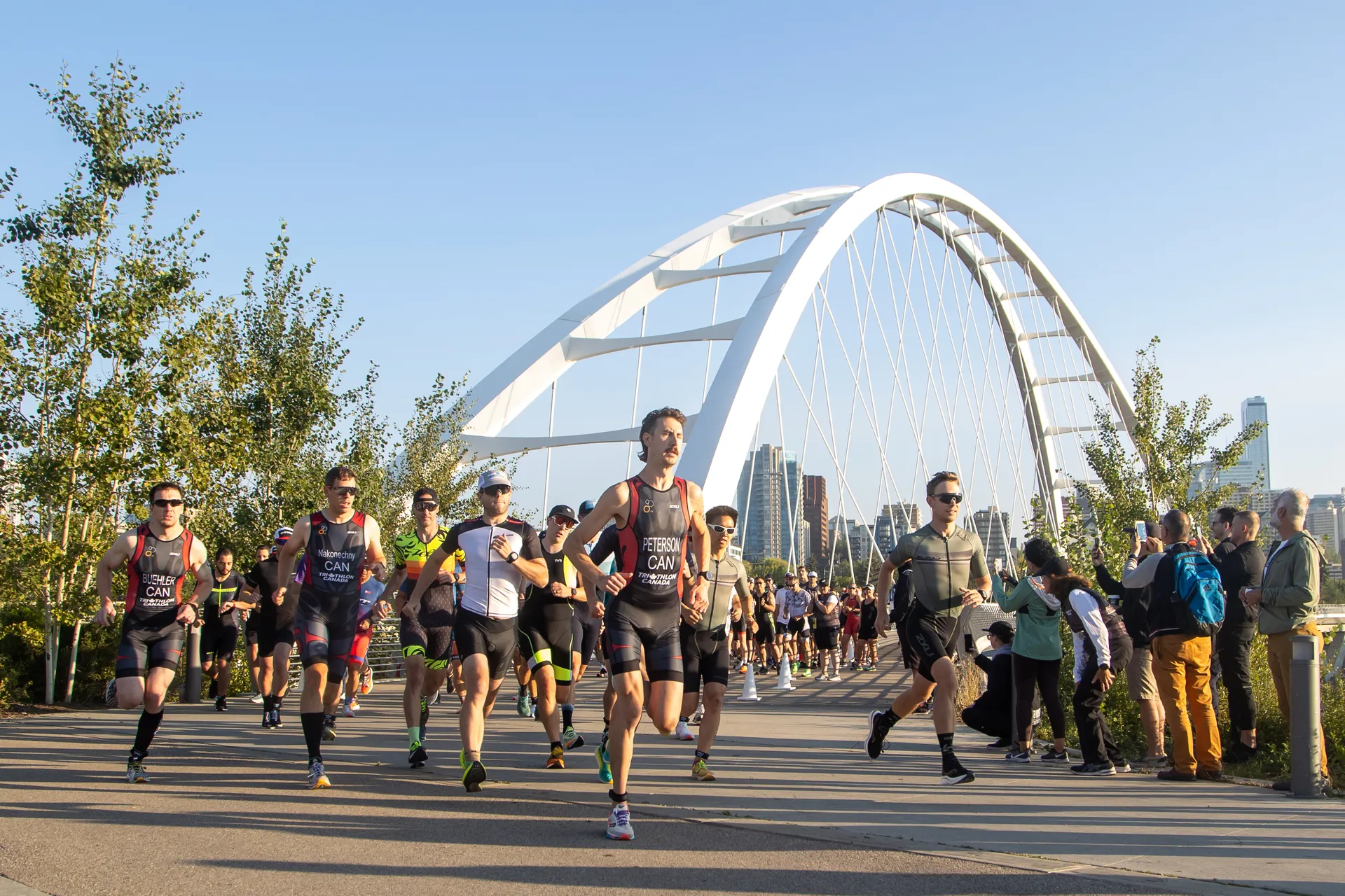The image shows a group of runners participating in a race or marathon. They are running on a path that appears to be a bridge or part of a park, with a distinctive white arch structure in the background. The runners are wearing athletic gear and race bibs, suggesting this is an organized event. Spectators are lining the side of the path, watching and possibly cheering on the participants. It's a sunny day, and the setting seems to be in an urban park, with tall buildings visible in the distance, indicating the event is likely taking place in a city.