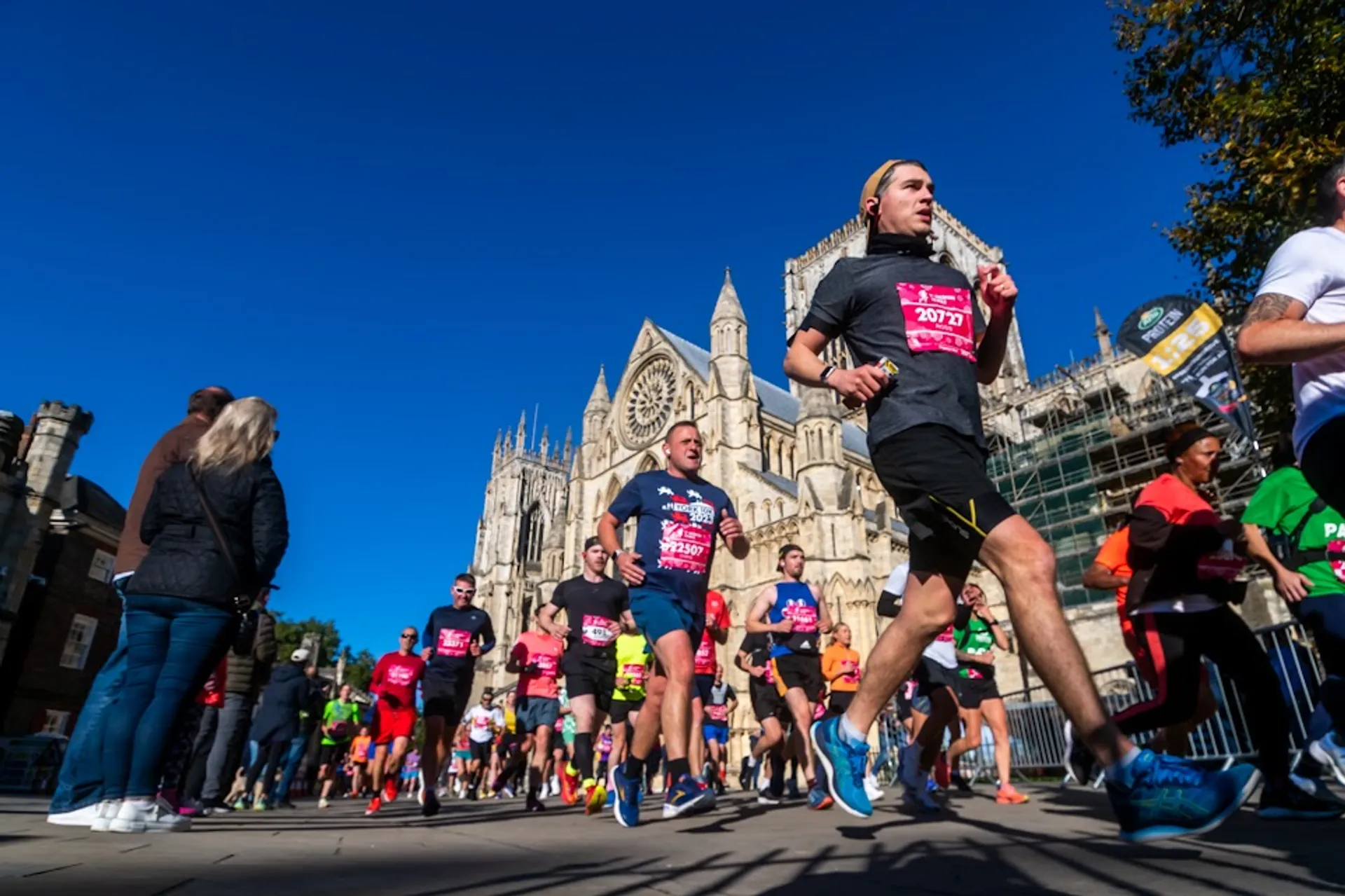 The image shows a group of people participating in a running event, likely a marathon or a race. The runners are wearing athletic attire and bib numbers, which is typical for organized running events. Some participants are focused and running at a steady pace, while others may be in the background. The background features a large, historic-looking building with Gothic architecture, suggesting that the event is taking place in a city with notable landmarks. The sky is clear and blue, indicating good weather conditions for the event. Spectators are visible on the side, suggesting it's a public, community event with local support and interest.