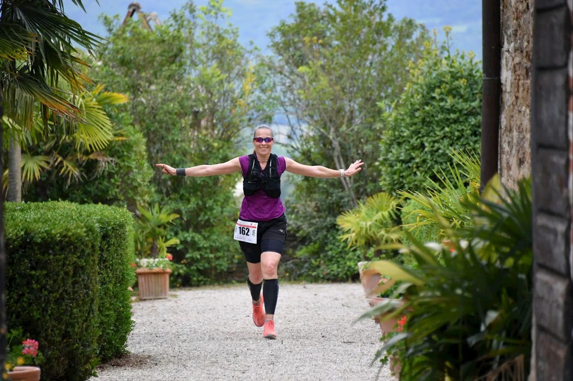 The image shows a person running outdoors along a gravel pathway, surrounded by lush greenery. The person is wearing a purple top, dark shorts, and has a race bib numbered 162. They appear to be happy, with arms outstretched, possibly finishing a race or enjoying the run. The background includes plants and stone structures.