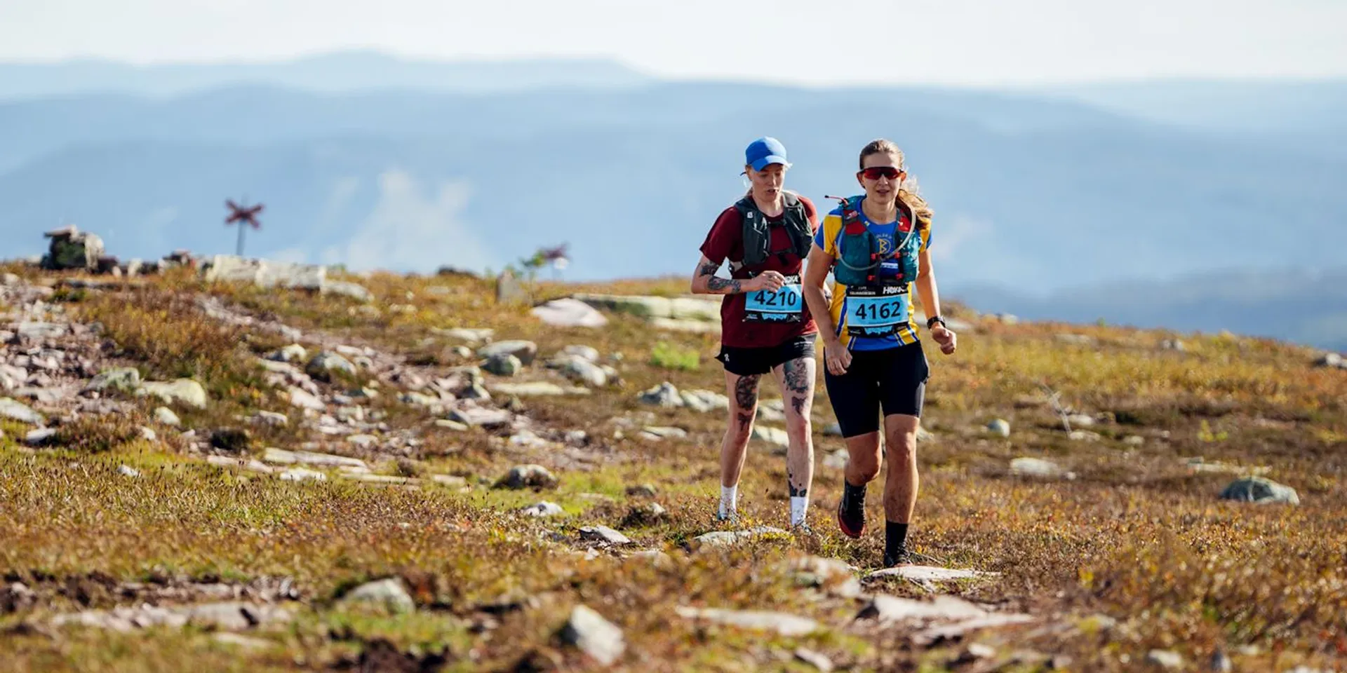The image shows two people running on a rocky, mountainous trail. They are wearing athletic gear and bib numbers, indicating they are likely participating in a trail running event or race. The background features expansive views of mountains and a clear sky.