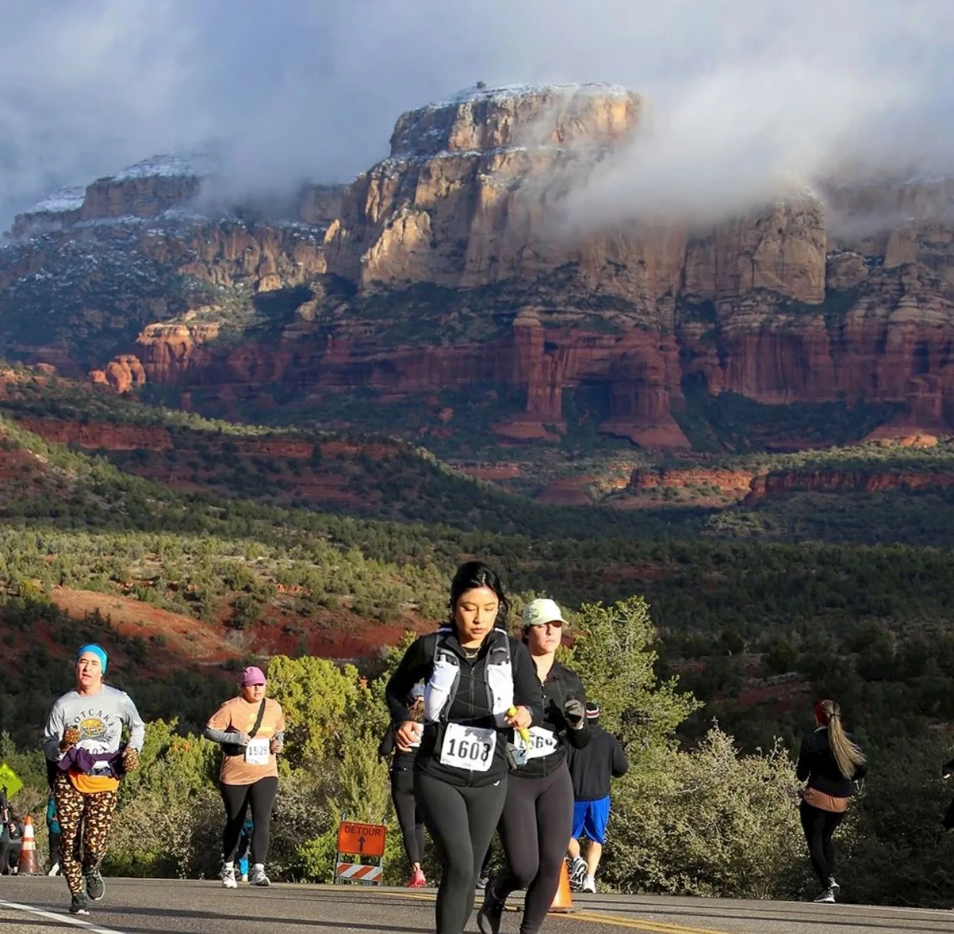 The image shows a group of people running in a scenic outdoor setting, likely participating in a race. The background features dramatic, rocky cliffs with some clouds or mist around them, suggesting it's a picturesque natural location. The runners are wearing athletic gear and race bibs.