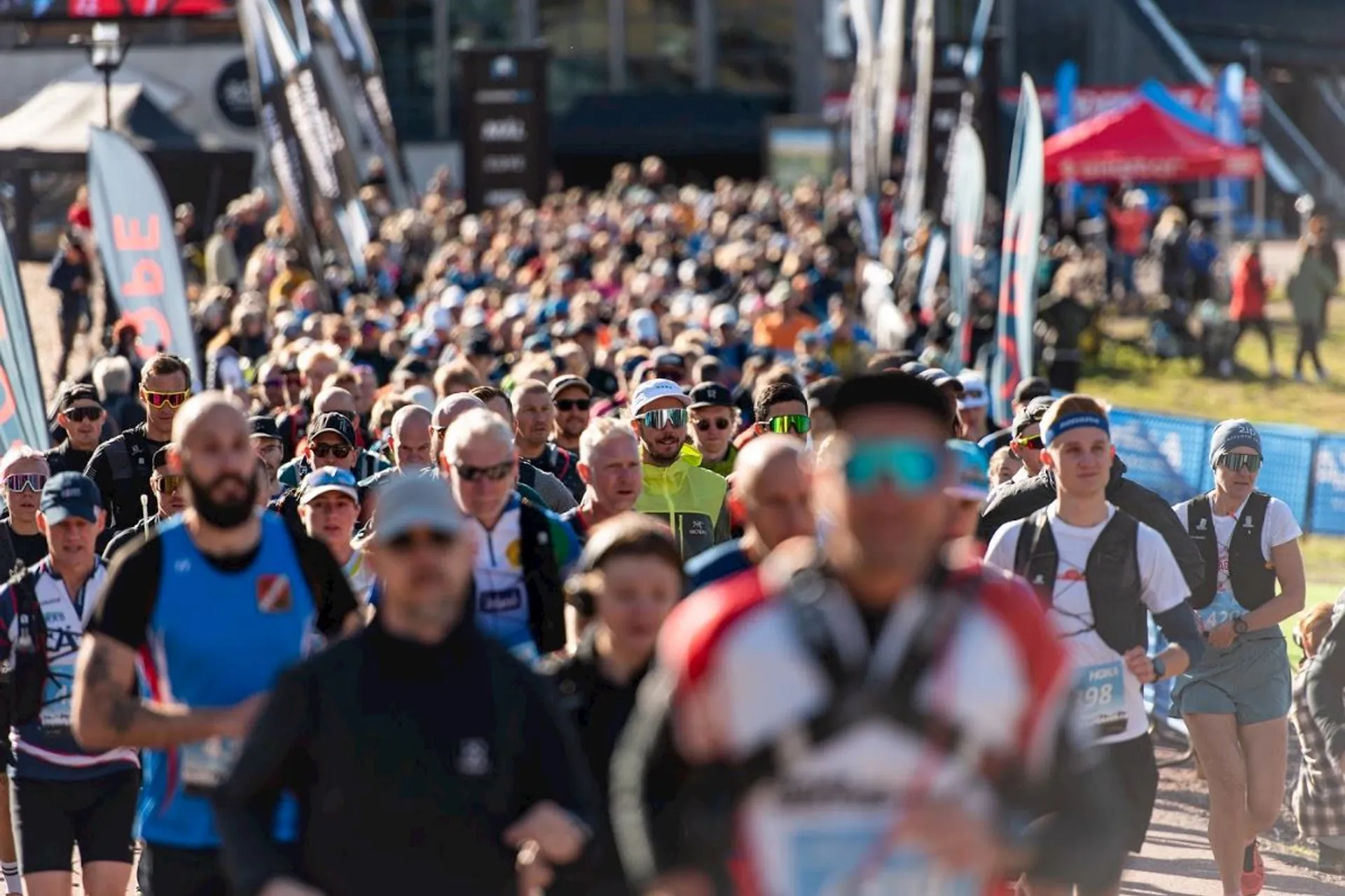 This image shows a large group of people participating in a running event. They are wearing athletic gear, such as hats, sunglasses, and running clothes. There are banners and tents in the background, suggesting an organized event.