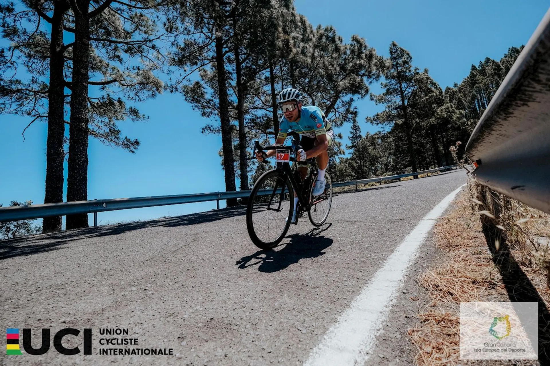 The image shows a cyclist riding aggressively up a mountain road during a cycling competition. The cyclist is wearing a number-themed bib, indicating they are part of a race or event. The environment suggests a sunny day with a clear blue sky. The road is bordered by guardrails on the right and dense trees can be seen in the background, showing that the road is likely in a forested or mountainous area. The logo in the bottom left corner indicates that the event is sanctioned by the UCI (Union Cycliste Internationale), the world governing body for sports cycling and international competitive cycling events. The logos and watermark suggest this image is likely from an official or professional source related to the cycling event.