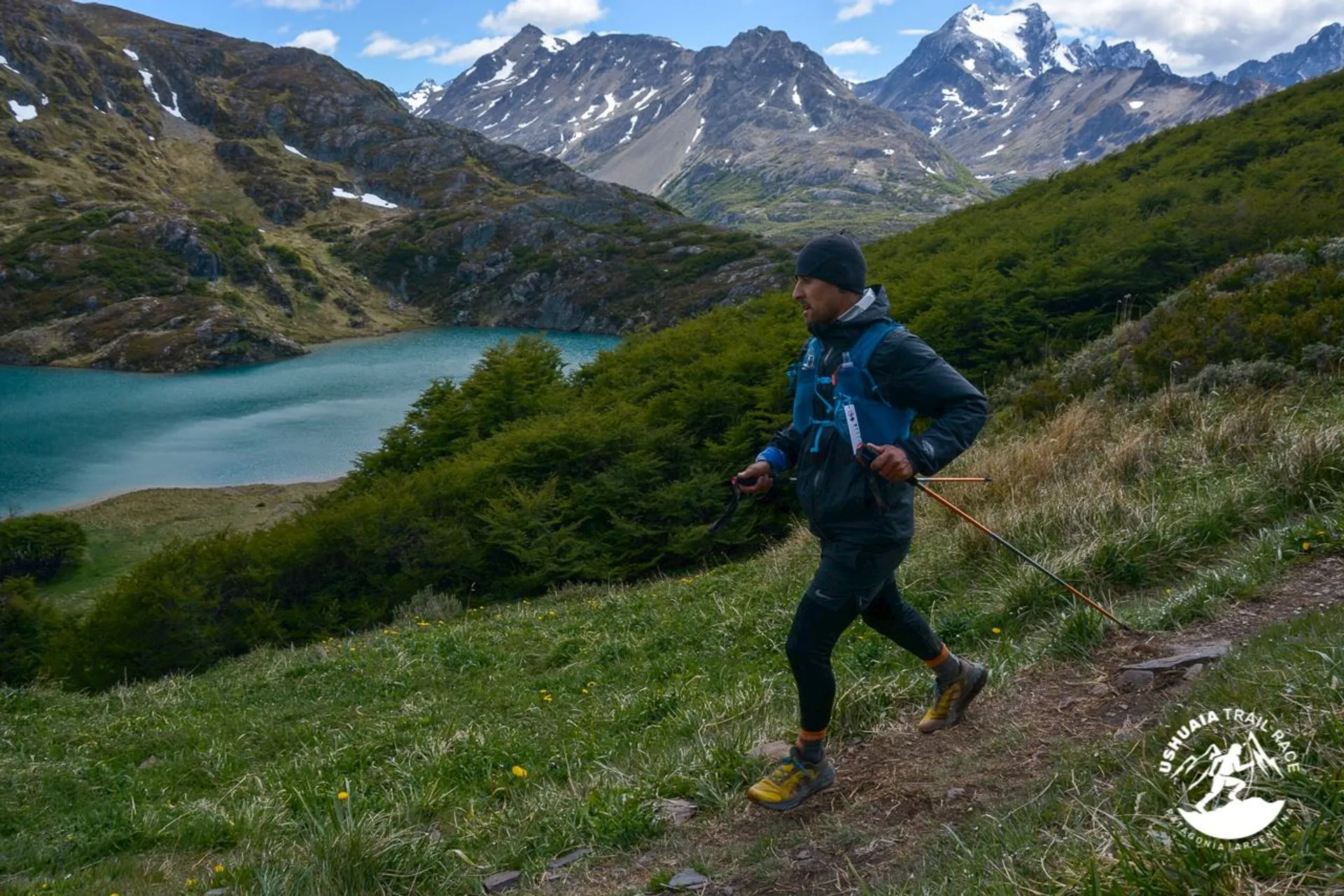 The image shows a person trail running in a scenic, mountainous landscape. There are snowy peaks in the background, a turquoise lake, and lush greenery. The runner is dressed in athletic gear and carrying poles, suggesting they are prepared for a challenging trail. The scene conveys a sense of adventure and connection with nature.