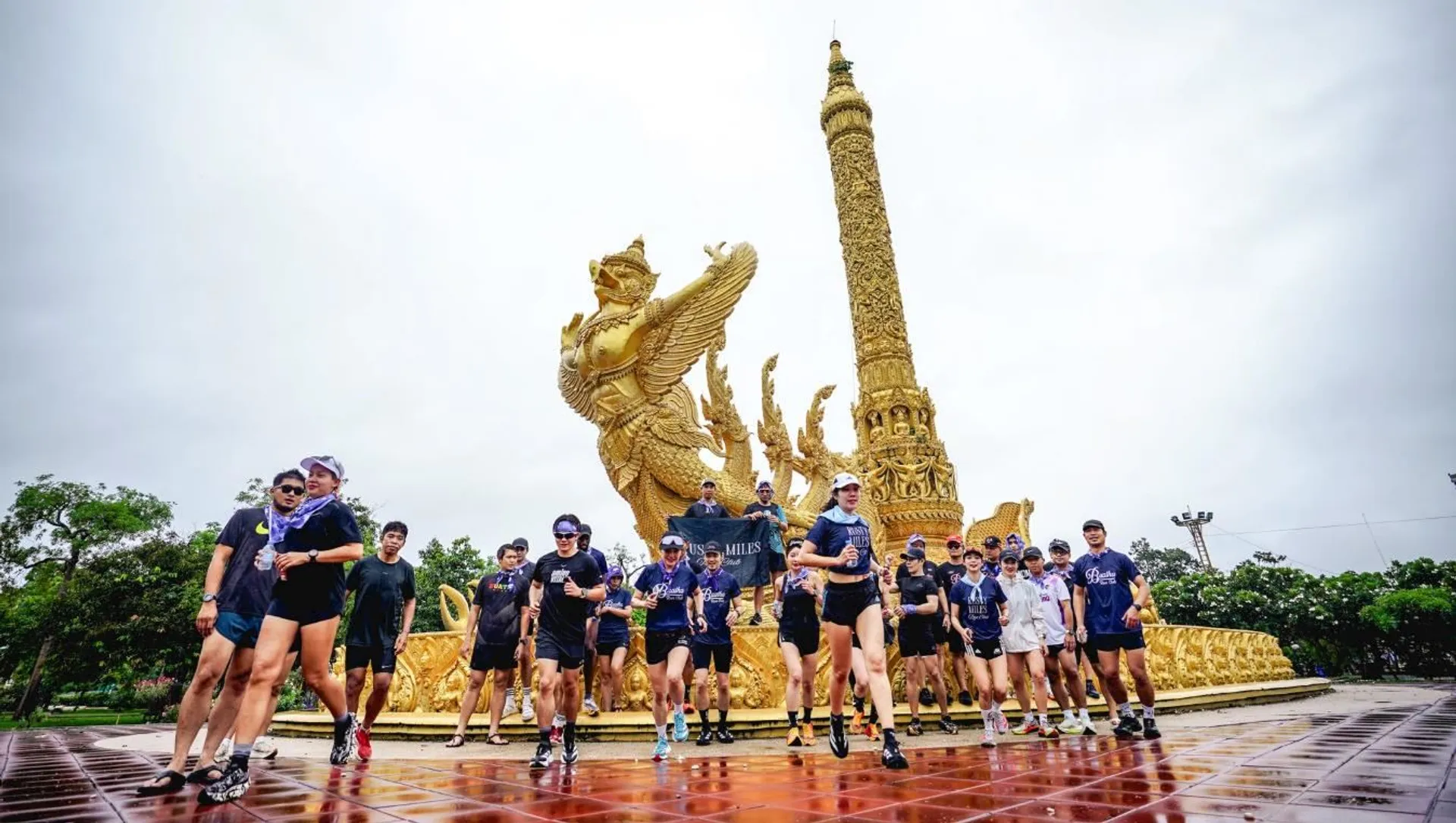 The image shows a group of people standing in front of a large, ornate golden statue with intricate designs. The statue includes a tall column and a mythical creature with wings. The group appears to be dressed in athletic or casual clothing. The location looks like it might be a public or cultural site, possibly in a park or plaza.