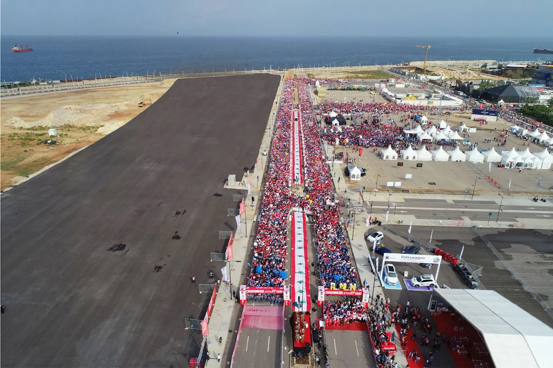 This image shows a large gathering of people in an outdoor event near the coast. There are tents and structures set up, suggesting it might be a festival or a similar event. The crowd is dense along a central pathway, and there are vehicles parked nearby. The event seems to have considerable attendance, and the area is organized with clear pathways and designated zones.