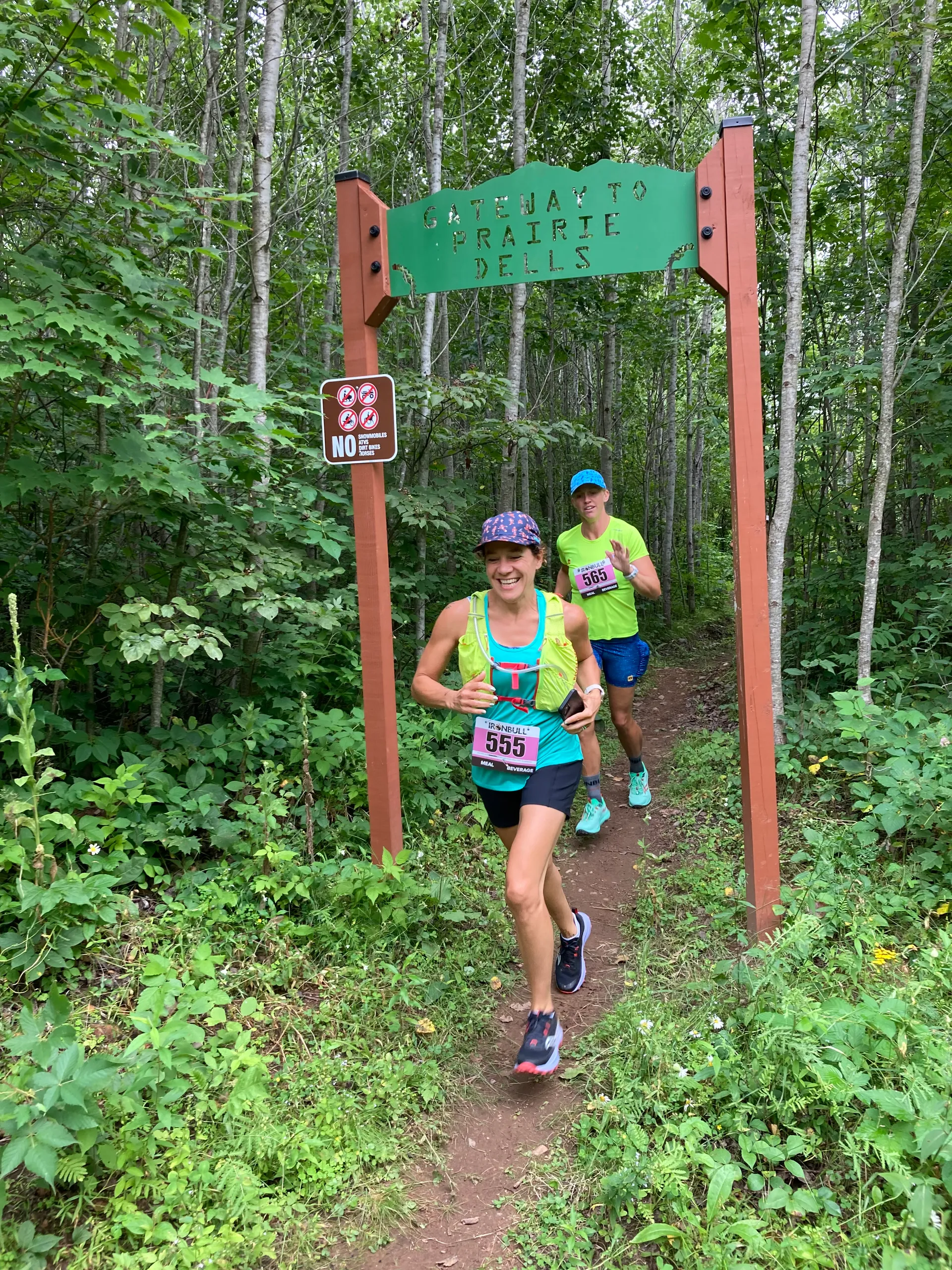 The image shows two people running on a forest trail under a sign that reads "Gateway to Prairies Dells." They are wearing athletic clothing and race bibs. The trail is surrounded by lush greenery, and a small sign nearby indicates that certain activities are not allowed.