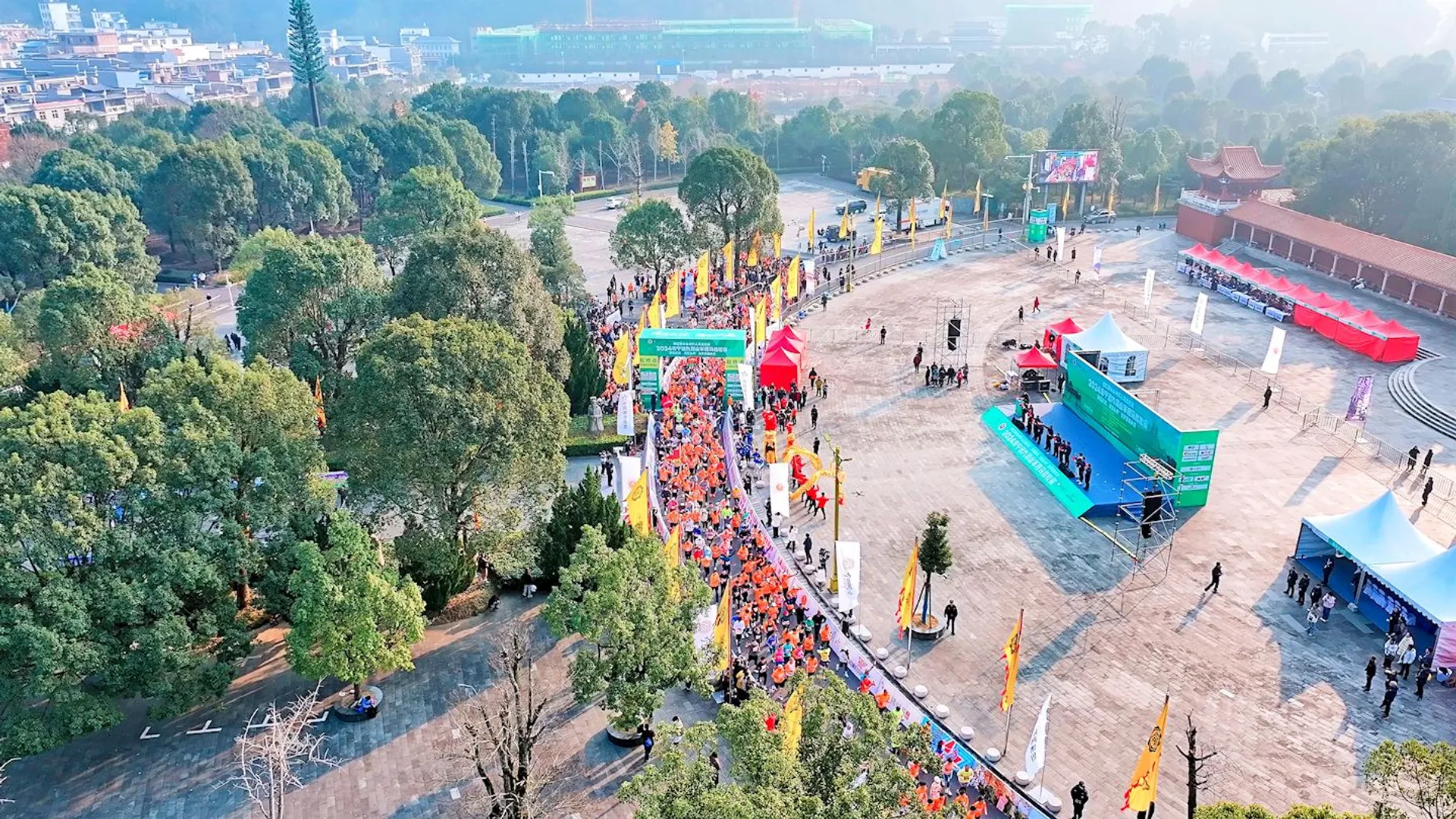 The image depicts a large outdoor event, possibly a race or marathon. There is a crowd of people, many wearing similar outfits, suggesting they might be participants in a run. Several tents, banners, and flags are set up, likely for event organization and promotions. The setting is a spacious area with trees and buildings in the background.