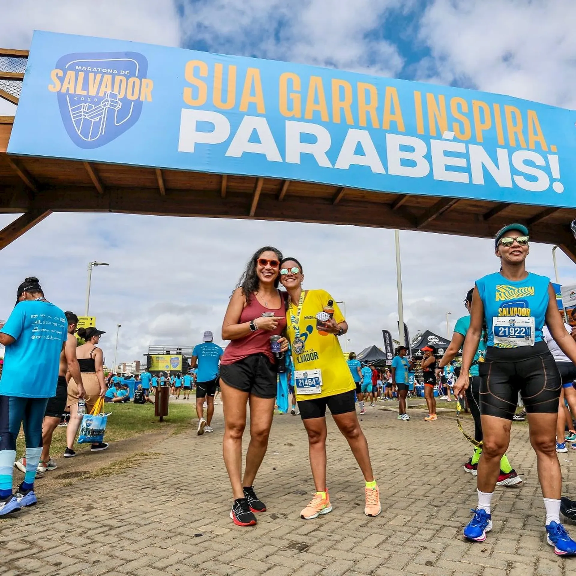The image depicts a group of people at a running event, with two women prominently featured in the foreground. They appear to be celebrating as they stand under a banner, which contains Portuguese text that translates to "Your grit inspires. Congratulations!" This indicates that they may have just finished a race and are receiving congratulations for their effort. The environment suggests a festive and athletic atmosphere, common to marathons or similar running events. Everyone is dressed in athletic wear suitable for running, and there are bib numbers visible on participants, which are typically worn by racers for identification. The location is indicated by the text on the participants' bibs and the banner as Salvador, which could refer to Salvador, Brazil, a city known for hosting various athletic events.