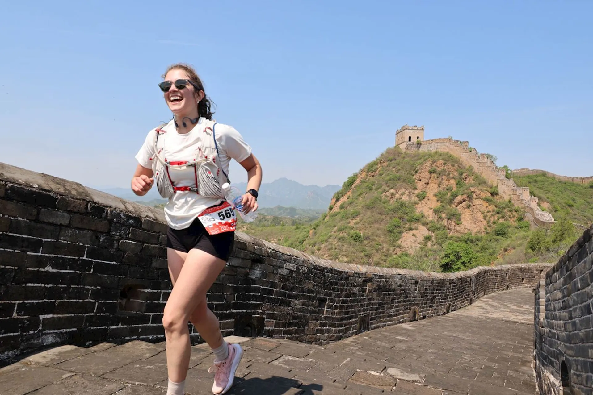 The image shows a person running on what appears to be the Great Wall of China. The person is wearing athletic clothing, sunglasses, and a hydration pack. There is a bib number on their shorts, suggesting participation in an event or race. In the background, there is a section of the Great Wall and natural scenery.
