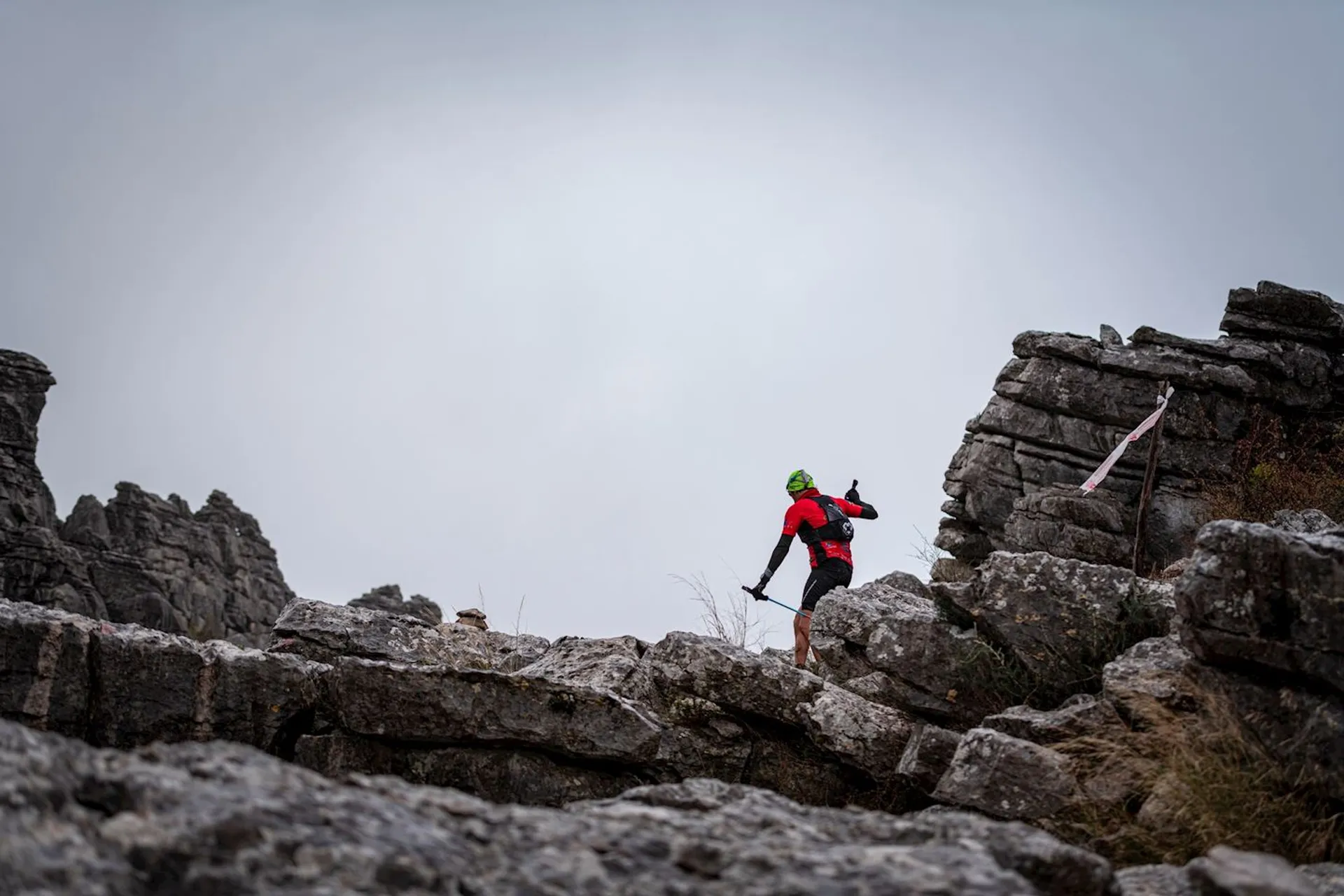 The image shows a person engaged in outdoor activity on rocky terrain. The person appears to be trail running or hiking and is dressed in cold-weather gear, suitable for the apparent chilly or possibly foggy environment indicated by the grey sky. They are wearing a red jacket with black pants and a green backpack, and they are in mid-stride, suggesting movement across the rough landscape. The rocks give the impression of a mountainous or high-altitude region. Markings that could be trail indicators are visible on some of the rocks, guiding the path for travelers.