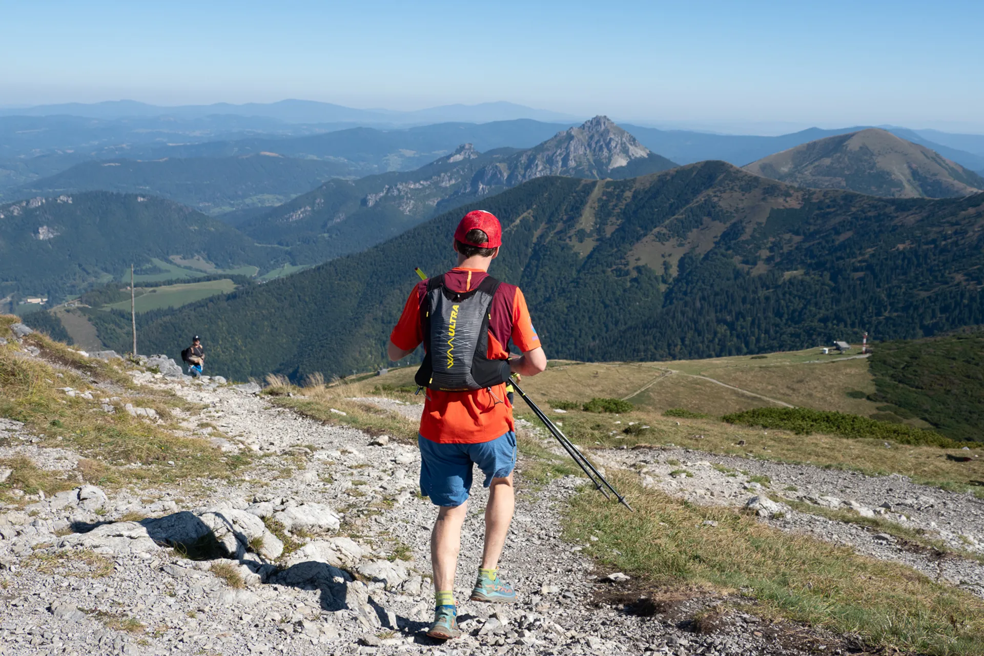 The image shows a person hiking on a rocky trail in a mountainous area. The hiker is wearing a red shirt, shorts, a backpack, and a red cap, and is holding trekking poles. The background features scenic views of mountains, valleys, and a clear blue sky.