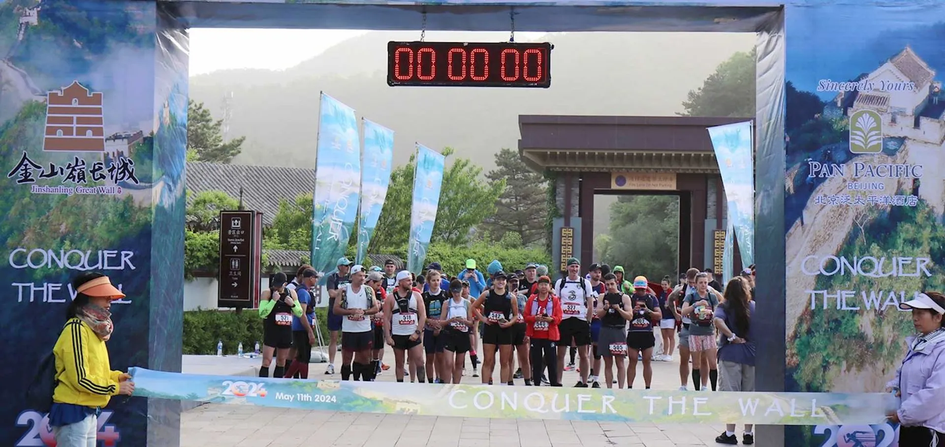 The image shows a group of runners at the starting line of a race called "Conquer the Wall." The event is associated with the Great Wall of China, as suggested by the entrance decor. There is a digital timer above the runners displaying "00:00:00" indicating that the race is about to start. The date on the banner is May 11th, 2024.