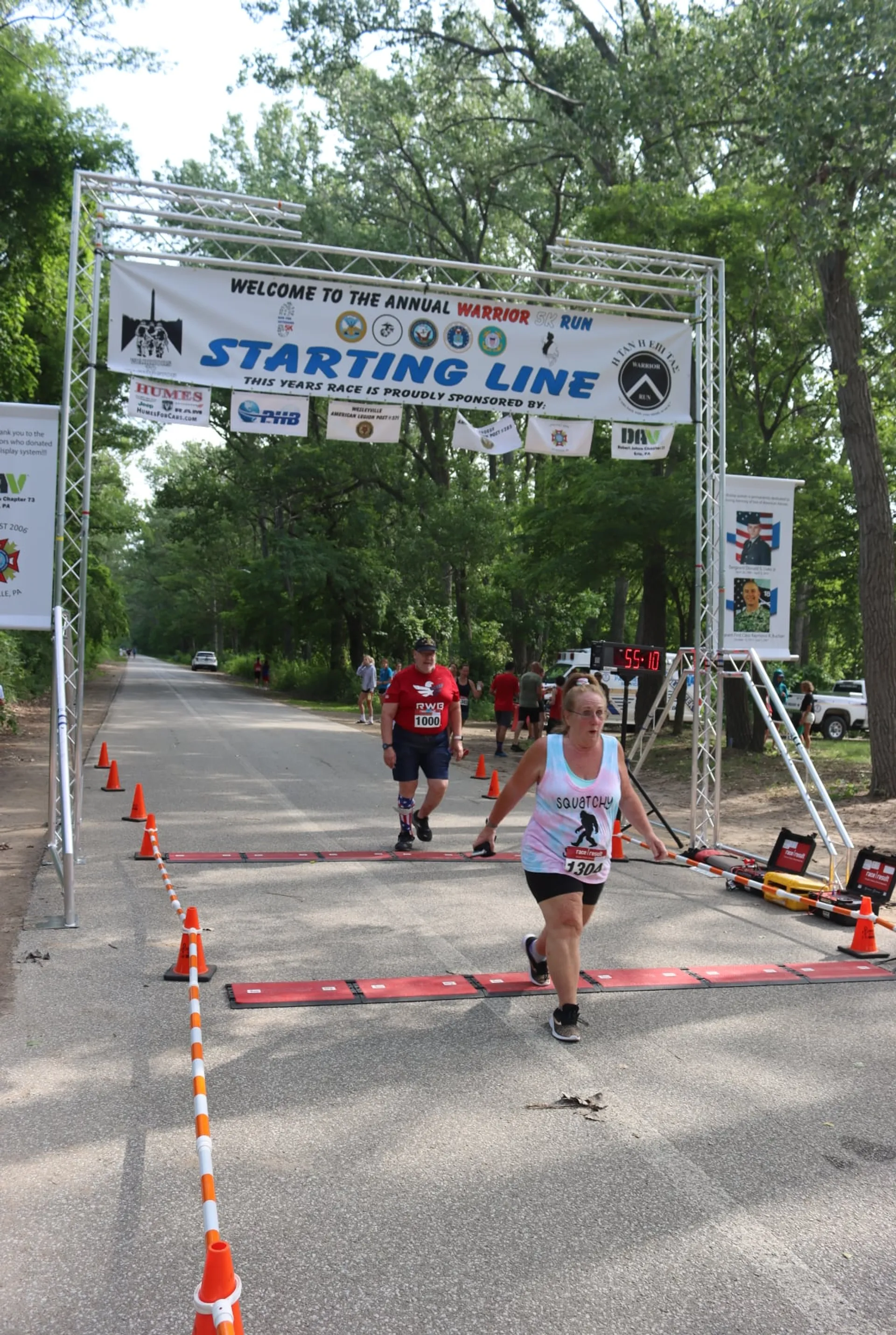 The image shows a running event where participants are crossing the starting line. There is a banner overhead that reads "WELCOME TO THE ANNUAL WARRIOR RUN STARTING LINE" with additional text that is not fully visible. Below the banner is the actual starting line with a timing mat on the ground where runners' times are likely being recorded electronically as they cross. We see a woman in the foreground wearing a number bib, indicating she is a participant in the event. She is in motion, suggesting that she has just begun her run. There are orange traffic cones lining the sides of the path, which seems to be in a park or wooded area, and another participant can be seen in the background. The environment is sunny and daylight, and