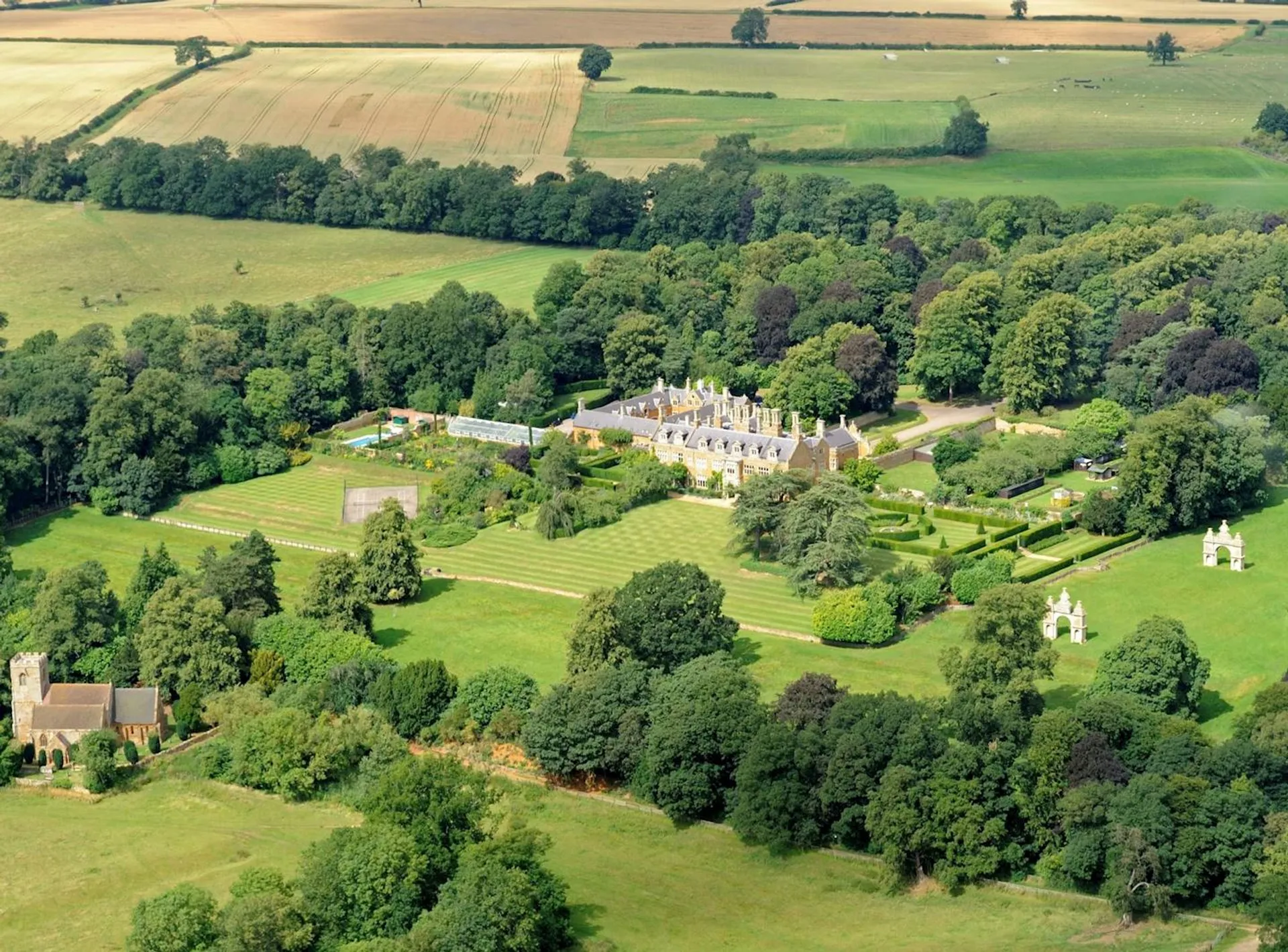 This image shows an aerial view of an expansive countryside estate. The estate includes a large mansion surrounded by well-manicured gardens and lawns. There are multiple hedges and pathways visible, along with several outbuildings. The area is surrounded by lush greenery, trees, and fields, providing a picturesque landscape typical of rural settings.