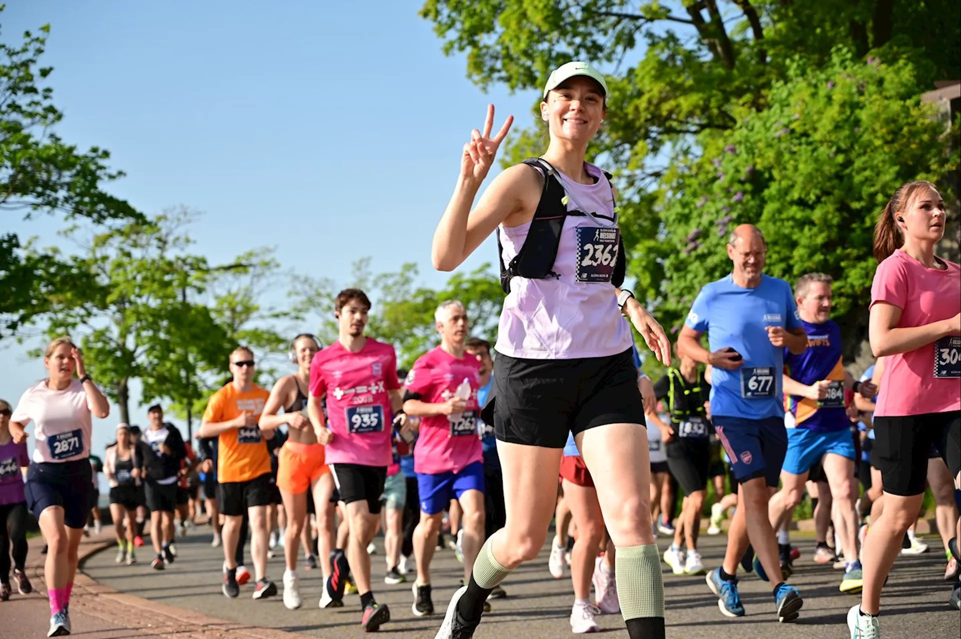 The image shows a group of people participating in a running event or marathon. They are wearing running gear with race numbers attached to their shirts. The setting is outdoors, with trees and clear skies visible in the background. One person is prominently displaying a peace sign and smiling at the camera.