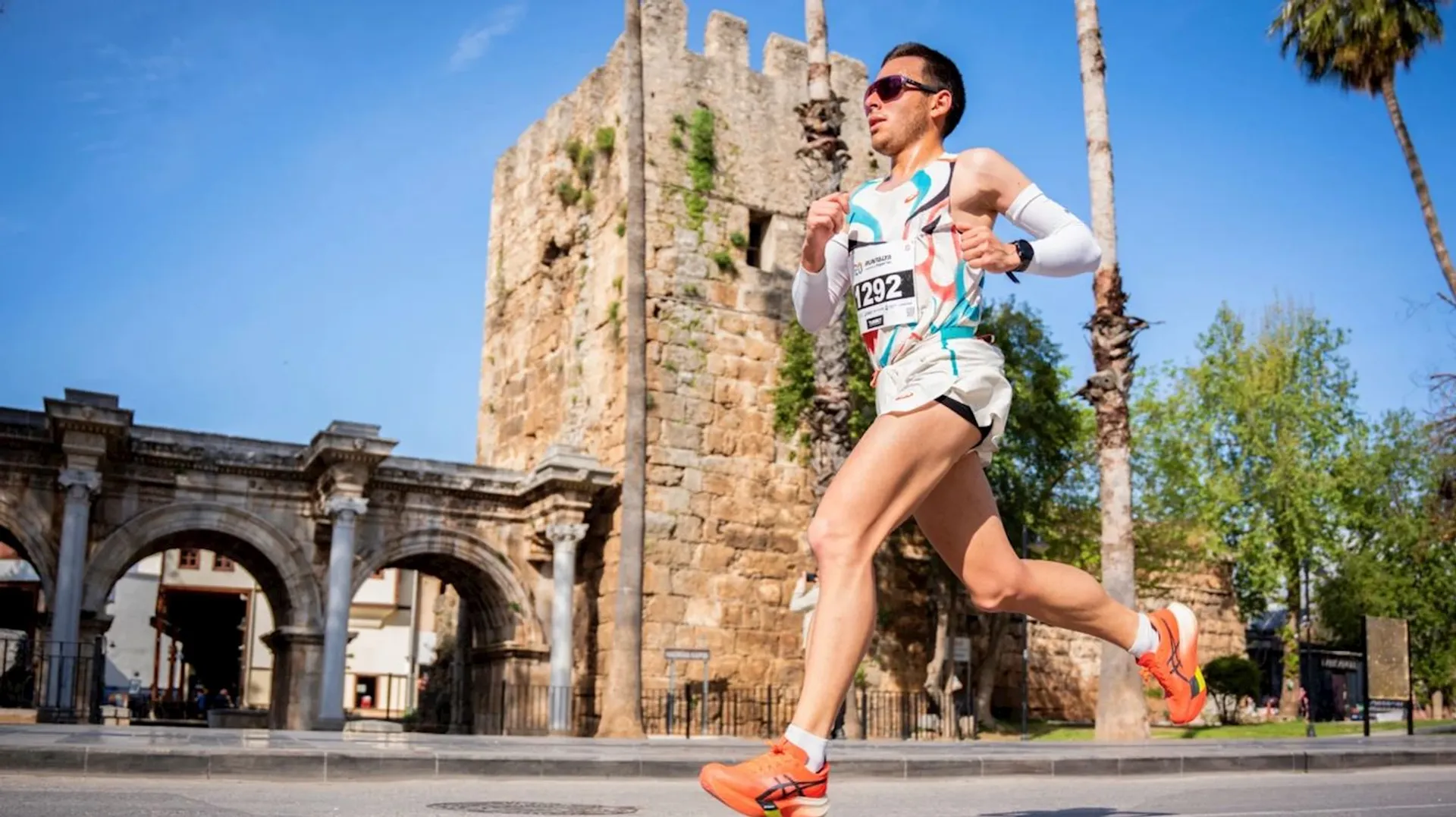 The image shows a person running in front of a historic stone structure, possibly a gate or wall, under a clear blue sky. The runner is wearing athletic gear and sunglasses, and there's a race bib visible on their outfit. Palm trees can be seen in the background.