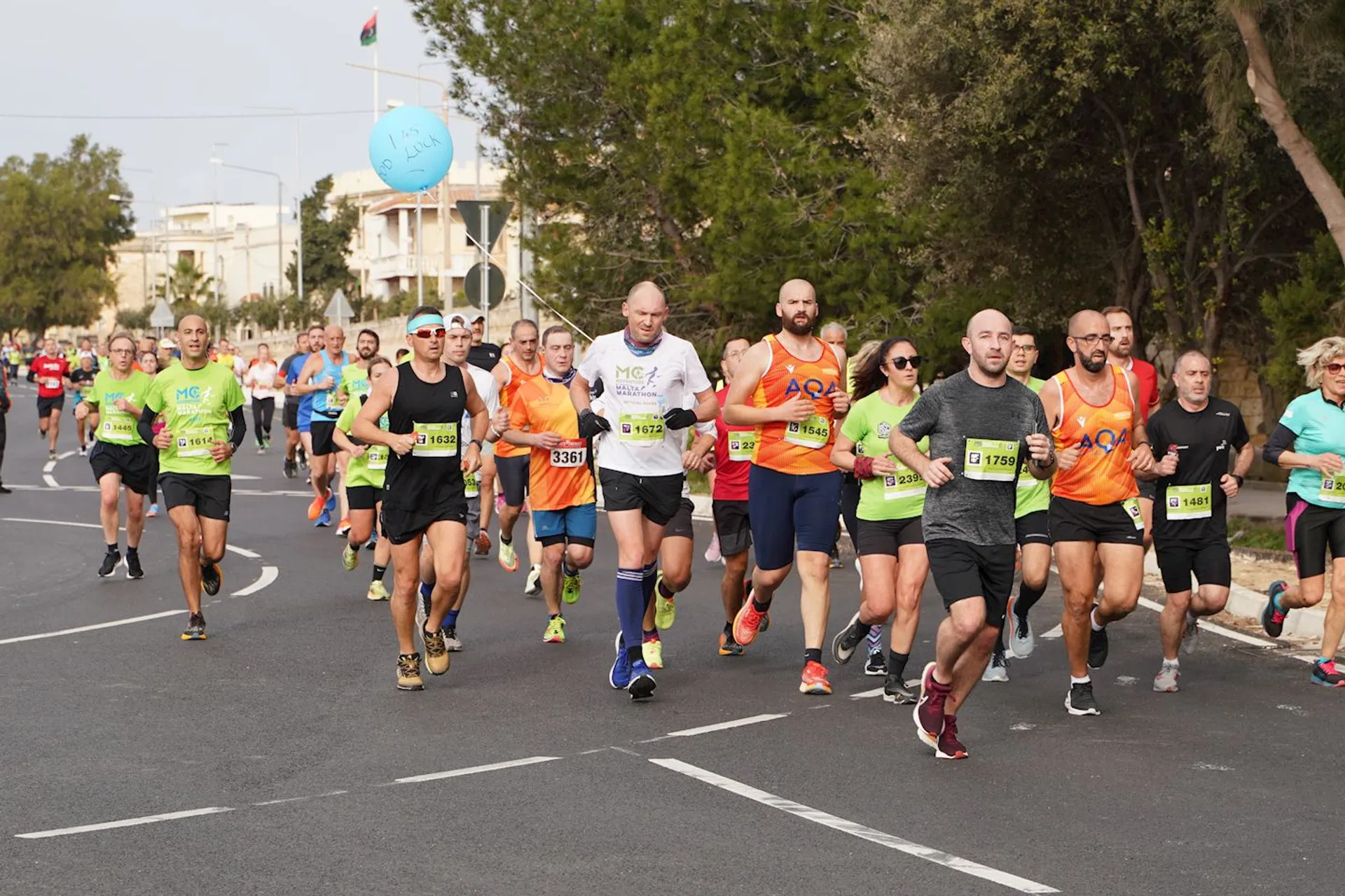 The image shows a group of people participating in a running event or marathon. They are running along a road, and some are wearing numbered race bibs and athletic clothing. Trees are visible in the background.
