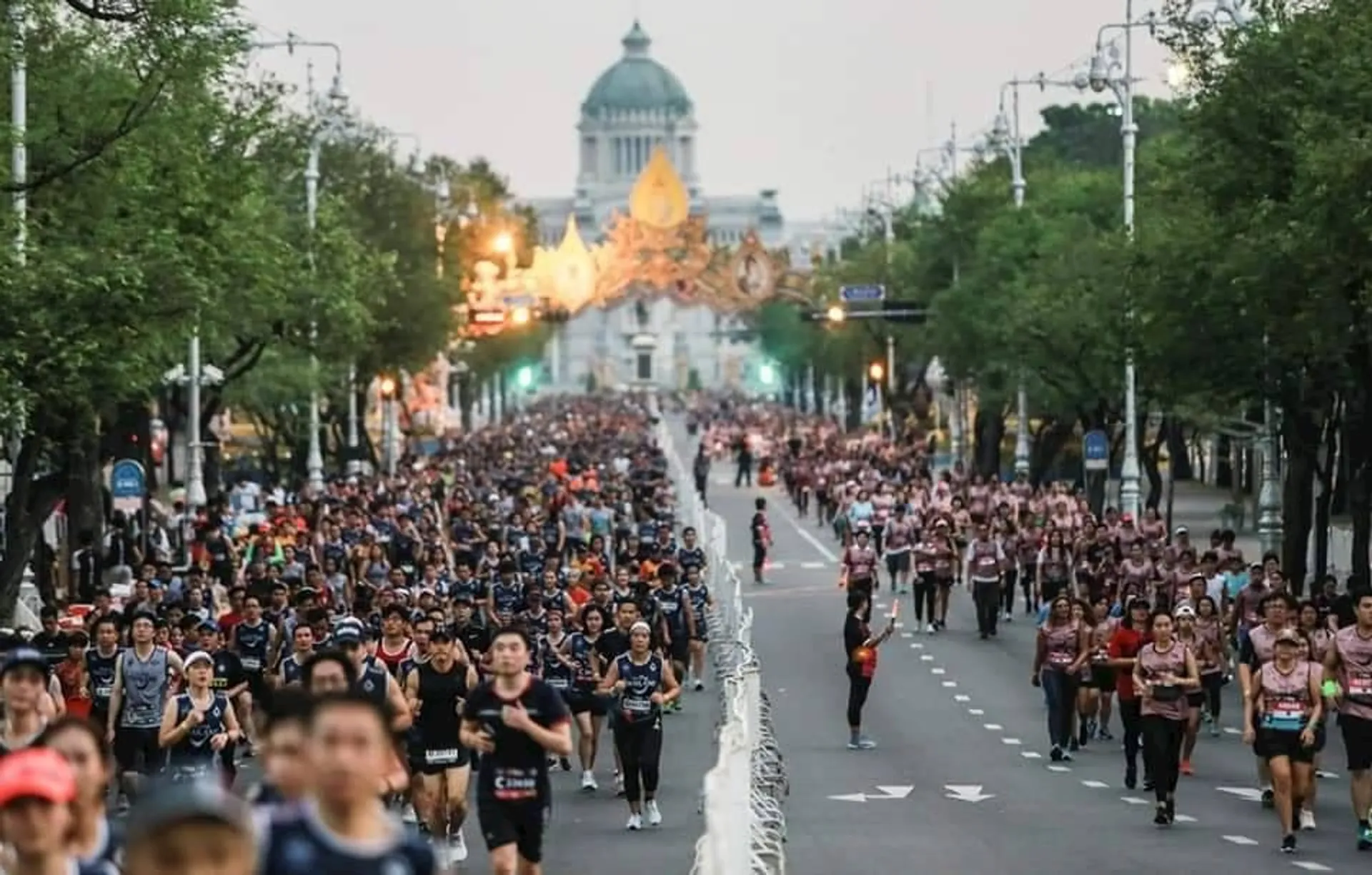 The image shows a large group of people participating in a marathon or running event on a wide street. The street is lined with trees, and there is a prominent building in the background. The atmosphere appears lively, with many participants wearing running gear.