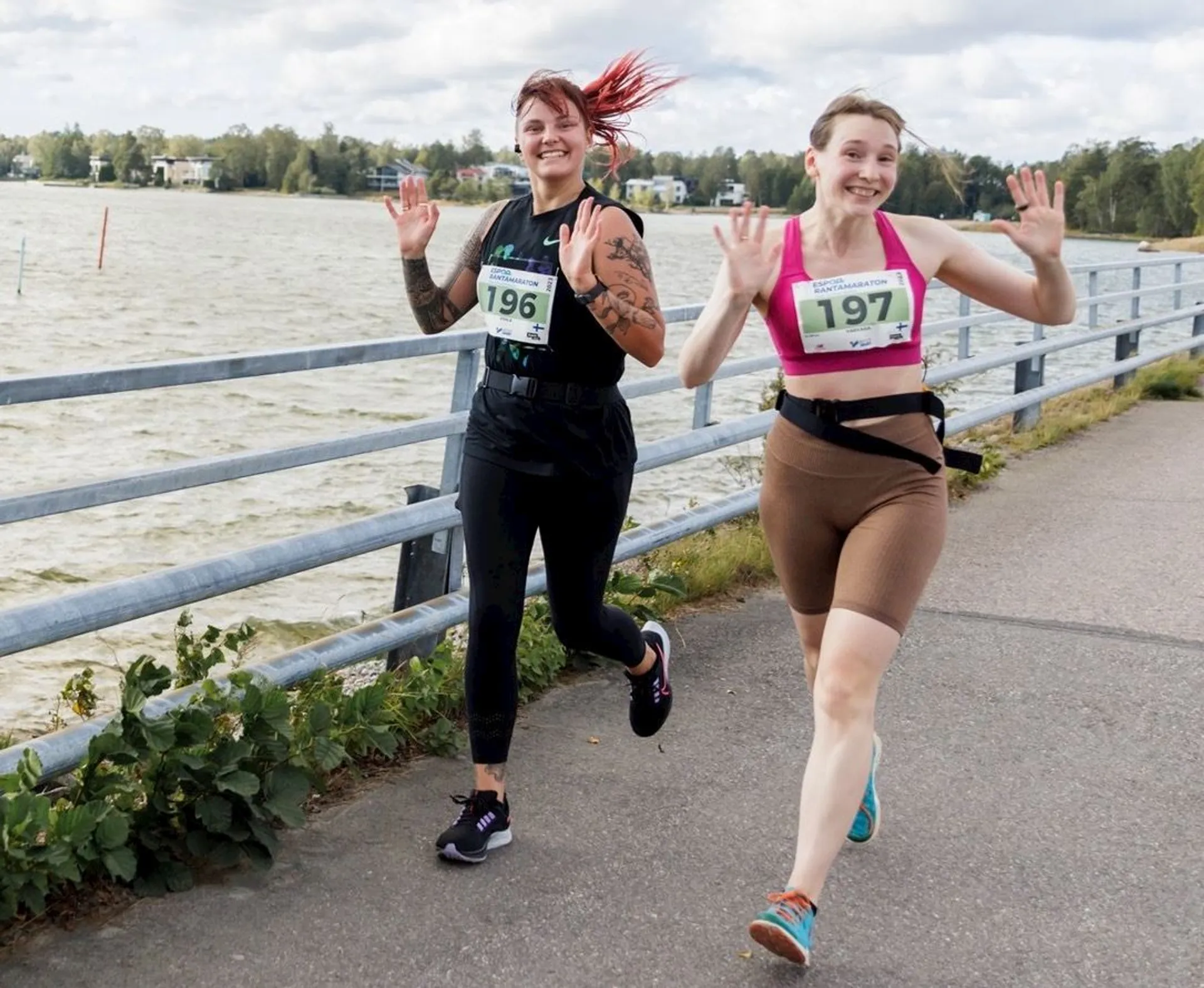 The image shows two people running on a path near a body of water. They are smiling and waving at the camera. Both are wearing numbers on their shirts, indicating they might be participating in a race or running event. The background features railing and some water, possibly a lake or river, with trees visible in the distance.