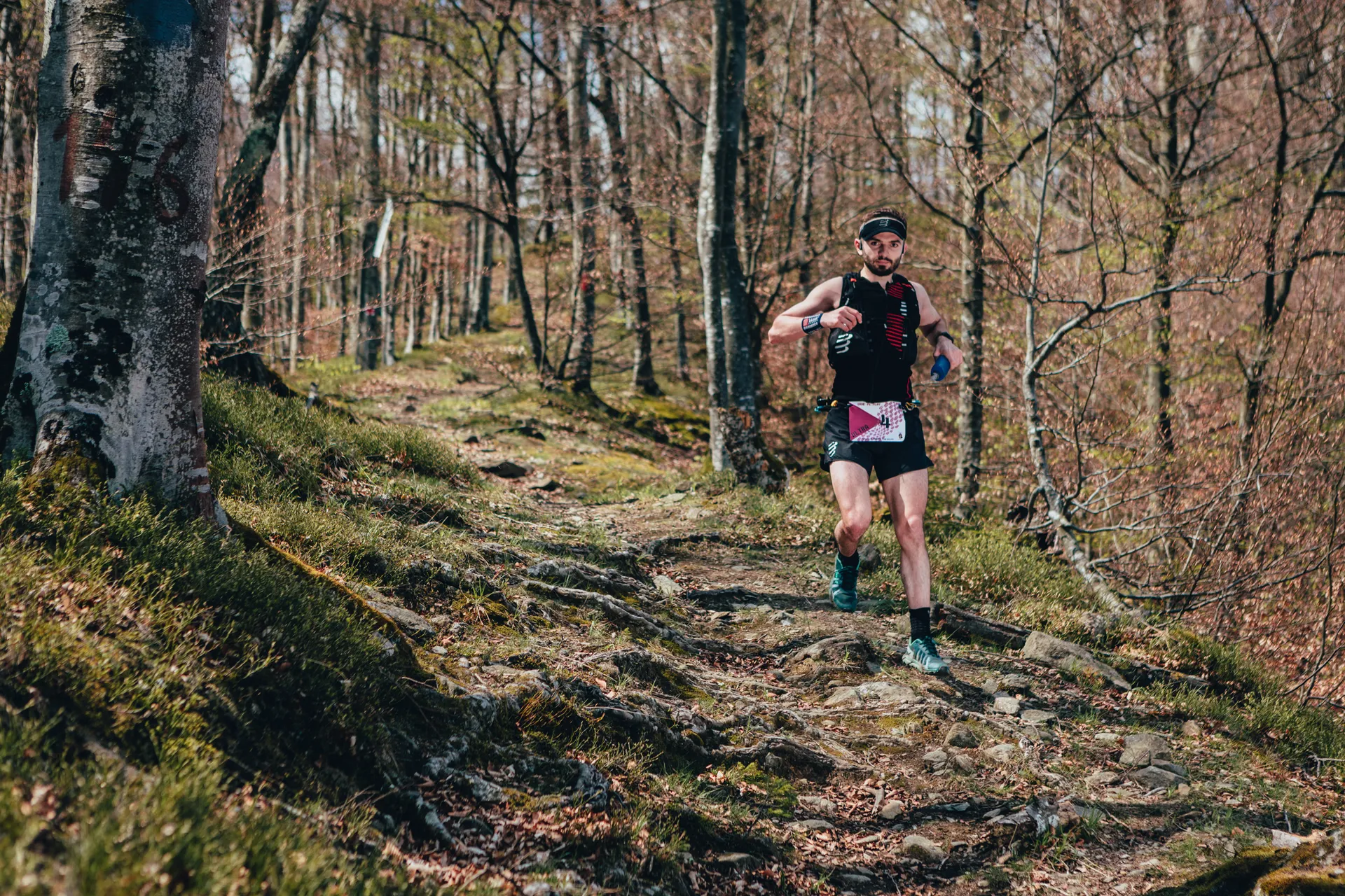 The image shows a person trail running through a forest. The runner is wearing athletic gear and appears to be on a dirt path surrounded by trees and natural scenery.