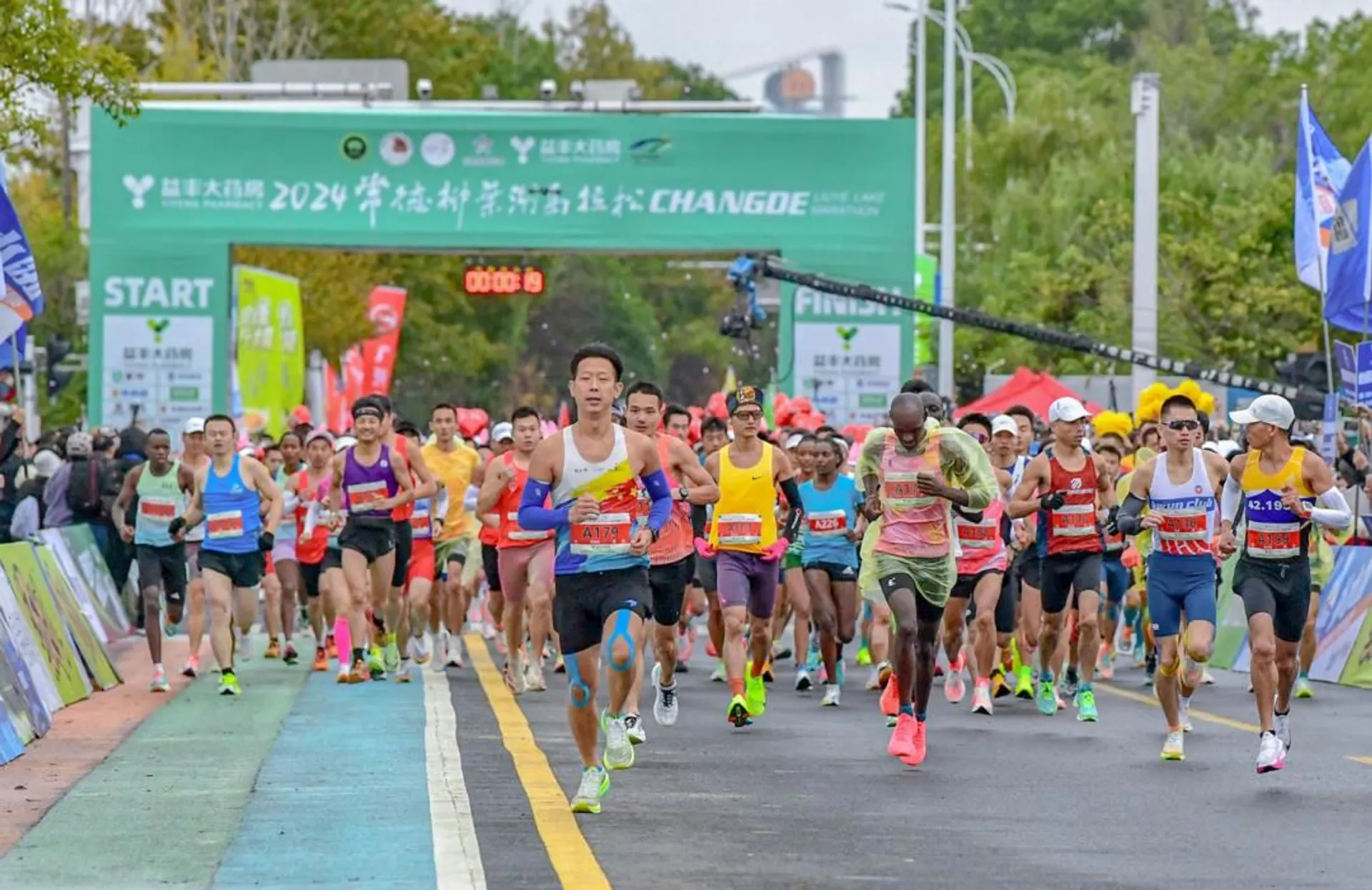 The image shows a group of runners participating in a marathon or race event. They are running on a marked course, heading towards the start/finish line. There are banners and signs around the area, indicating it is a formal event. The setting appears to be outdoors, with spectators in the background.