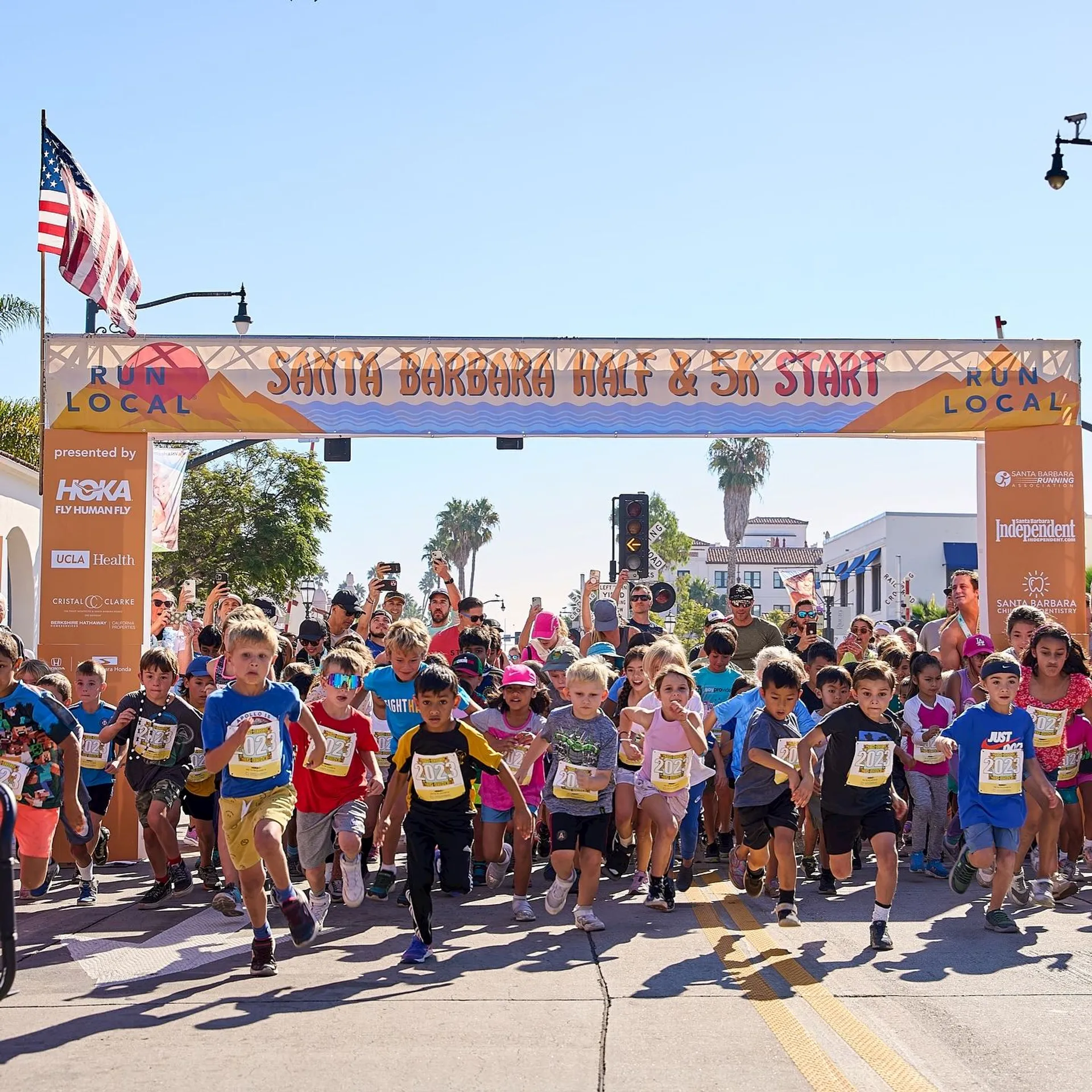 The image depicts the start of a race event called "Santa Barbara Half & 5K." A large group of runners, including children, is seen at the starting line. An American flag and several sponsor banners are visible in the background. The atmosphere is lively, suggesting a community event.