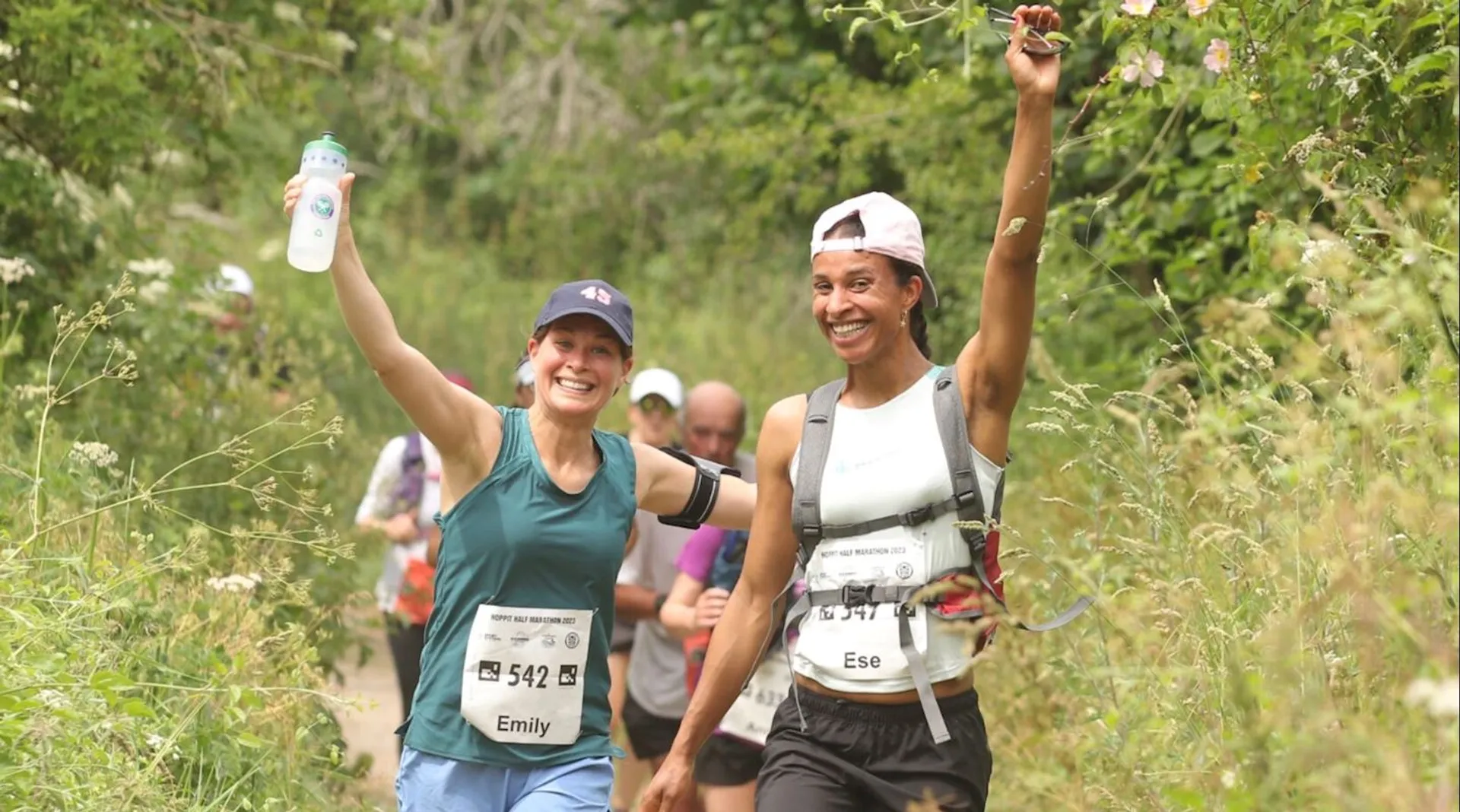 The image shows two people participating in a running event. They are smiling and appear to be in a natural, outdoor setting with greenery around them. Both are wearing athletic gear and have race numbers on their fronts. One of them is holding a water bottle and raising it in a celebratory manner.