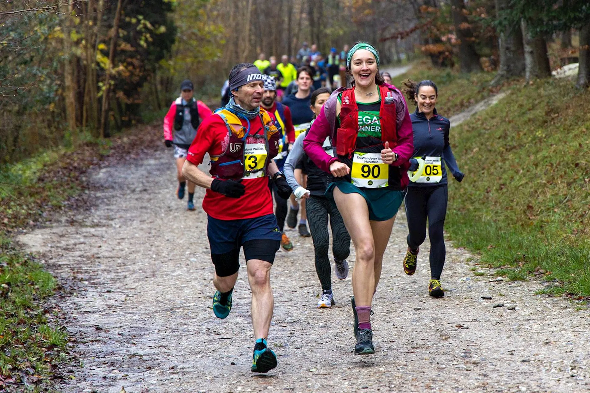 The image depicts a group of people engaged in a running event, possibly a trail run, as they are running on a dirt path surrounded by trees indicating a natural, outdoor setting. The participants are wearing athletic clothing and bib numbers, which indicate that this is an organized race. They seem to be in good spirits, with one runner smiling and looking directly at the camera, giving the photo a dynamic and lively feel. The image captures a moment of physical activity, wellness, and possibly competition.