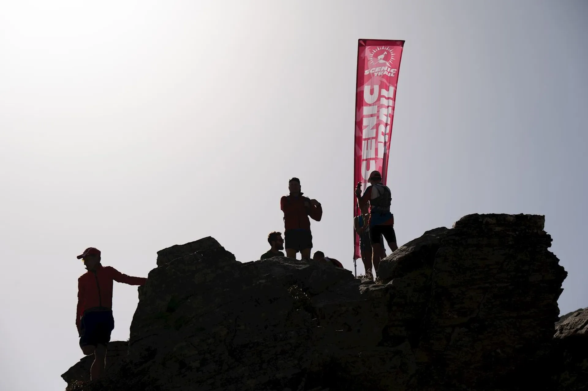 The image shows several people standing on rocky terrain. They are silhouetted against the sky, and one of them is holding a vertical red banner with text on it. The light suggests it may be sunrise or sunset, creating a striking silhouette effect.