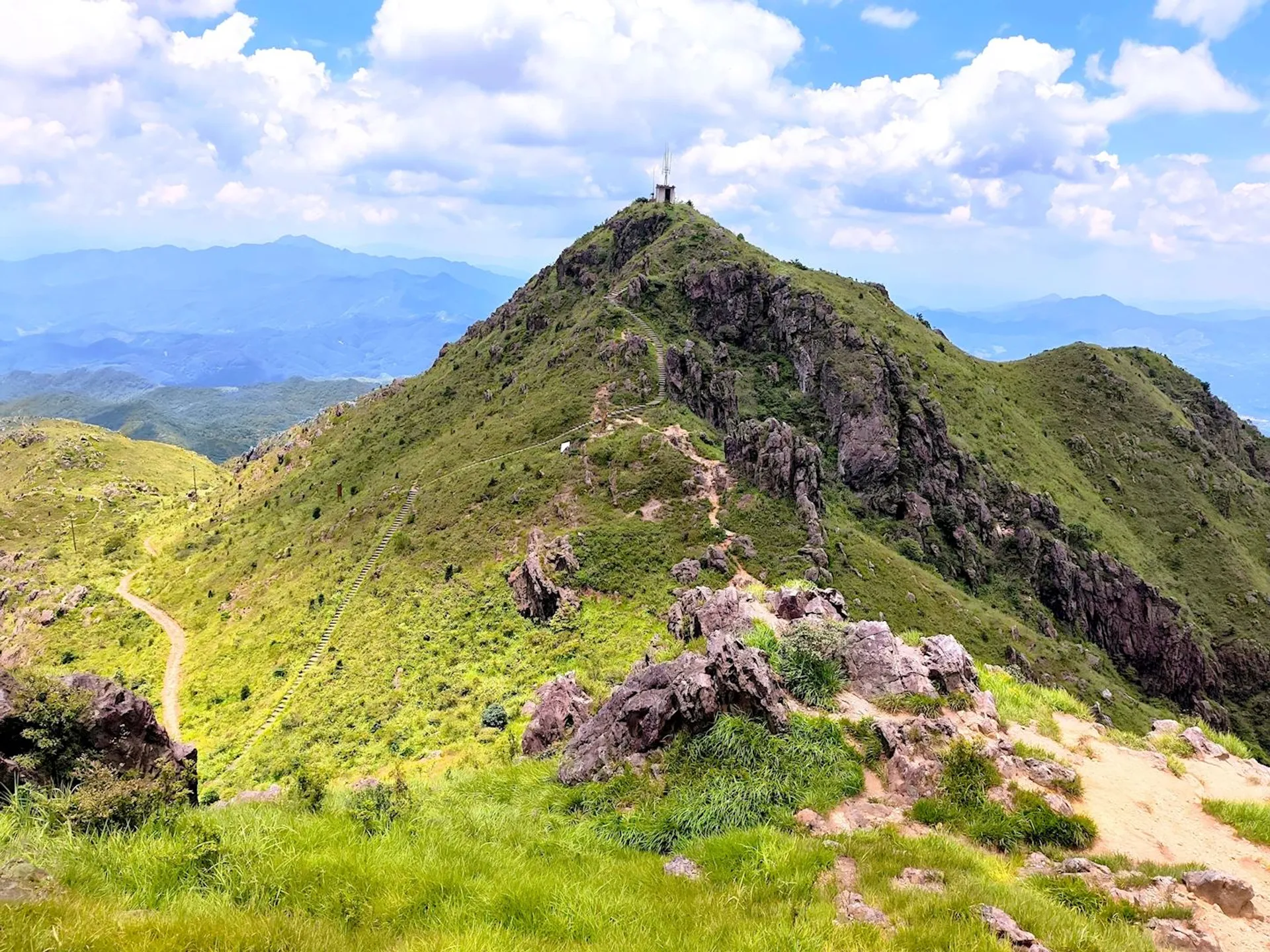 The image shows a scenic view of a mountain landscape. A grassy trail leads up to the peak, which is covered in green vegetation and has rocky outcrops. There is a small structure at the summit. The sky is partly cloudy, and mountains can be seen in the distance.