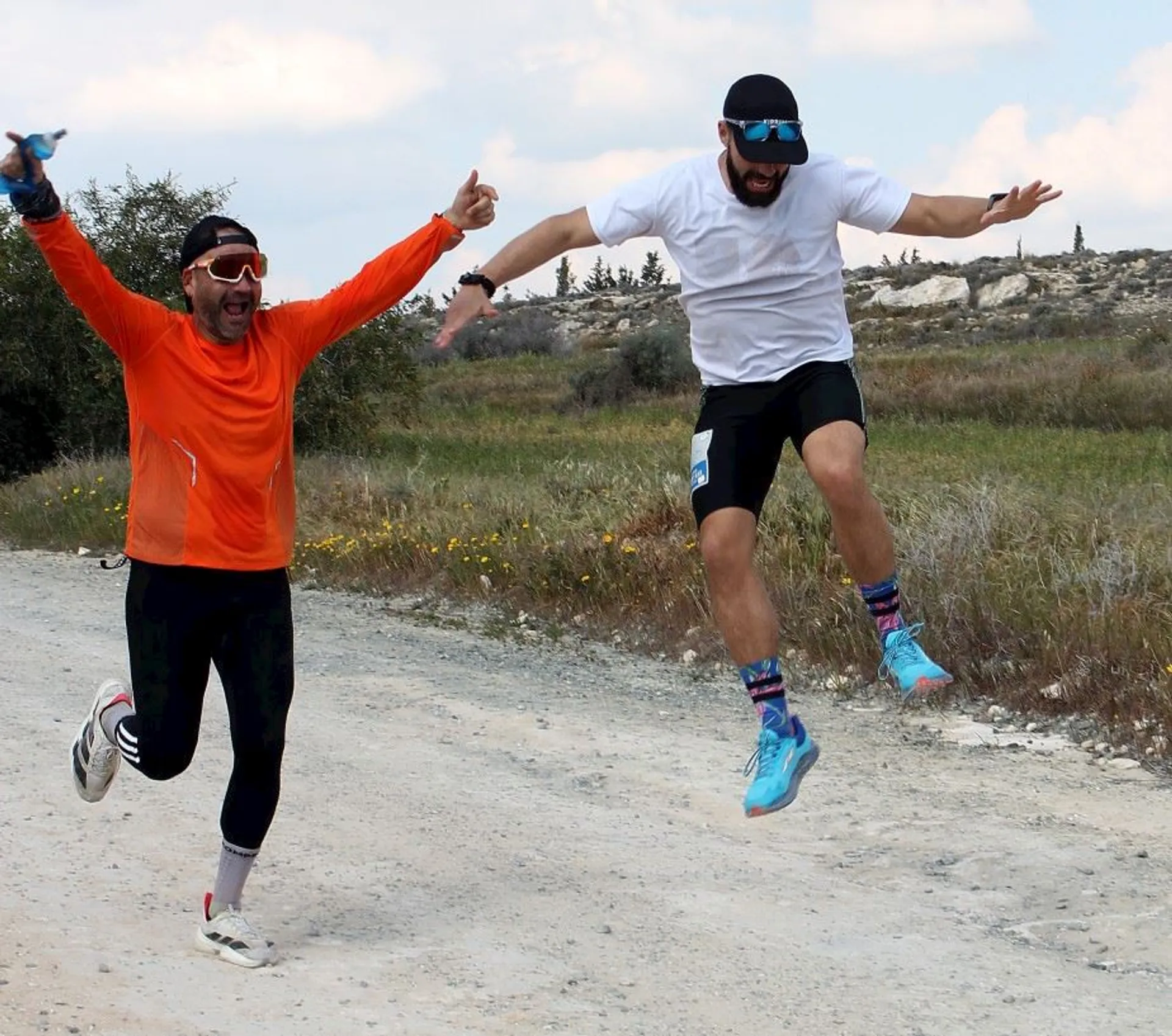 The image shows two people running outdoors on a dirt path. One is wearing an orange shirt and black pants, and the other is in a white shirt and black shorts. They appear to be jumping with their arms raised, looking joyful. Both are wearing caps and sunglasses. The background includes grass and trees under a partly cloudy sky.