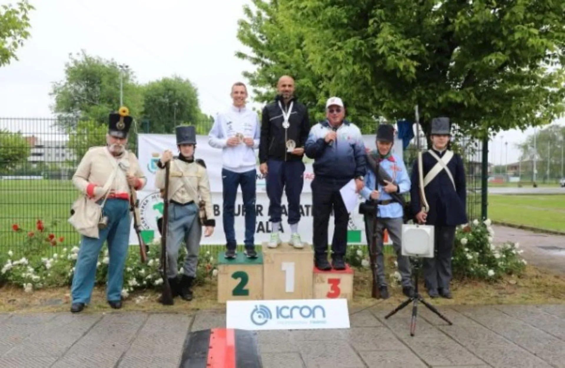 The image shows a group of people standing on a winner's podium during what appears to be an awards ceremony. There are three individuals on the podium itself, each standing on a different level indicating their respective positions in a competition—first, second, and third place. The person in first place is standing on the highest level, with a medal around their neck, and is in the center, while those in second and third places are on the lower levels on the left and right, respectively, also with medals.

Additionally, there are individuals standing on each side of the podium, appearing to be dressed in historical military uniforms, possibly re-enactors or part of a ceremonial event. The setting looks like an outdoor area, and there are some flowers and a